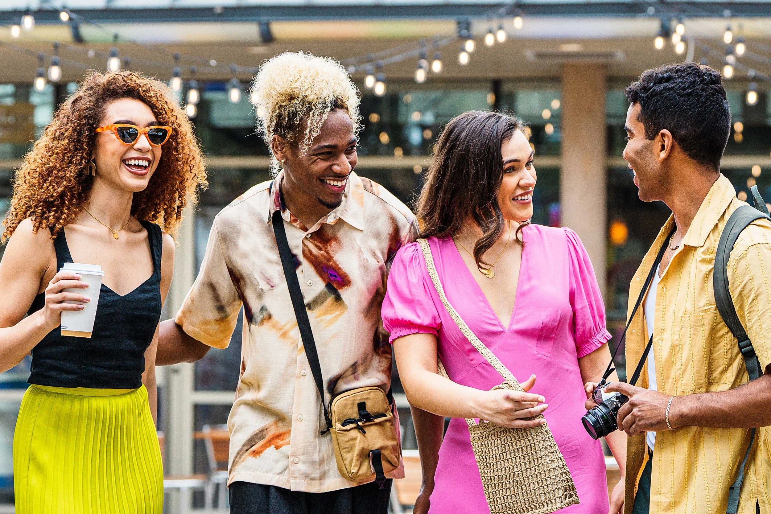 Four friends standing and talking outside, smiling and enjoying themselves, with string lights overhead.
