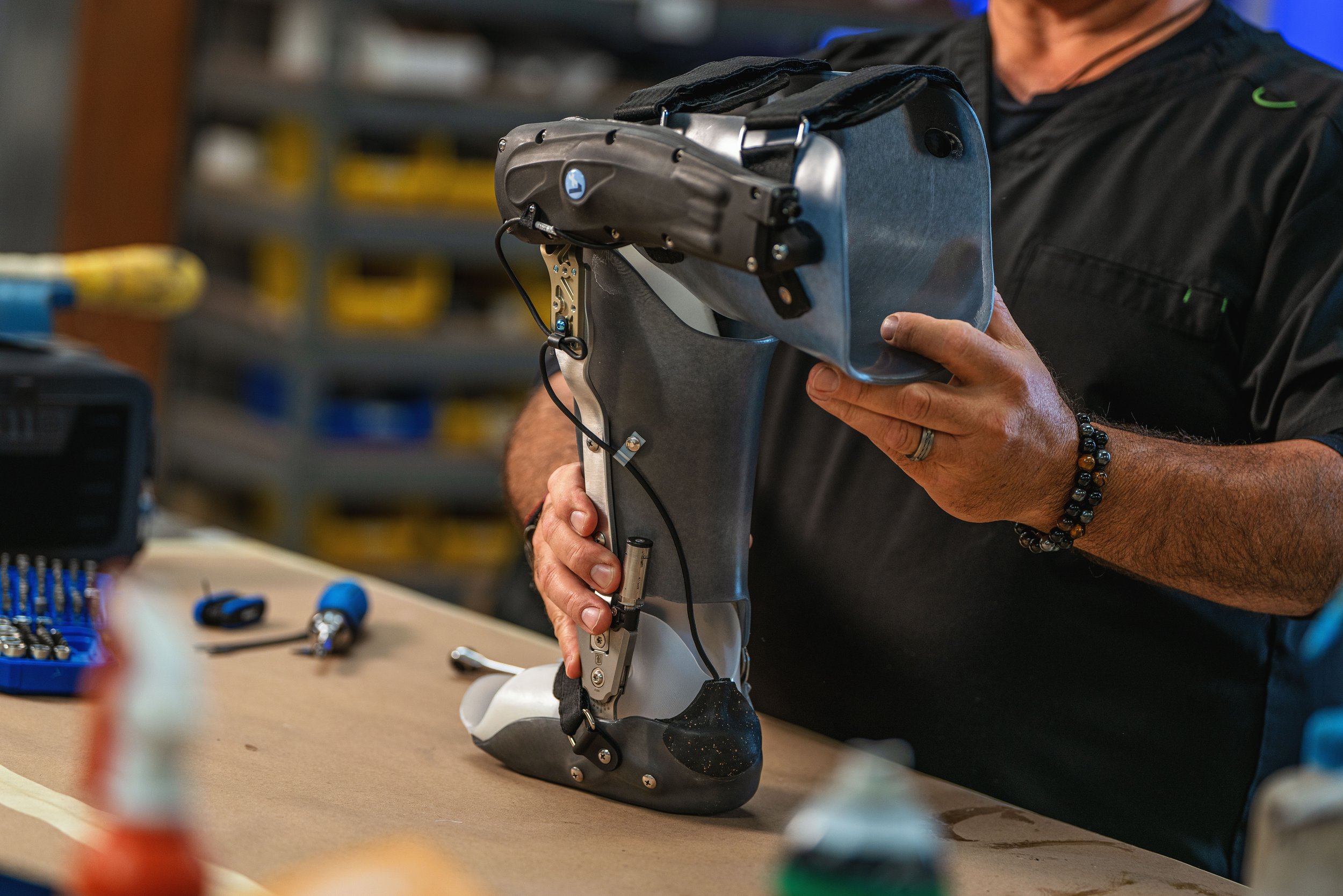 A person assembling or repairing a prosthetic leg in a workshop, holding the prosthesis with both hands, surrounded by tools and equipment.