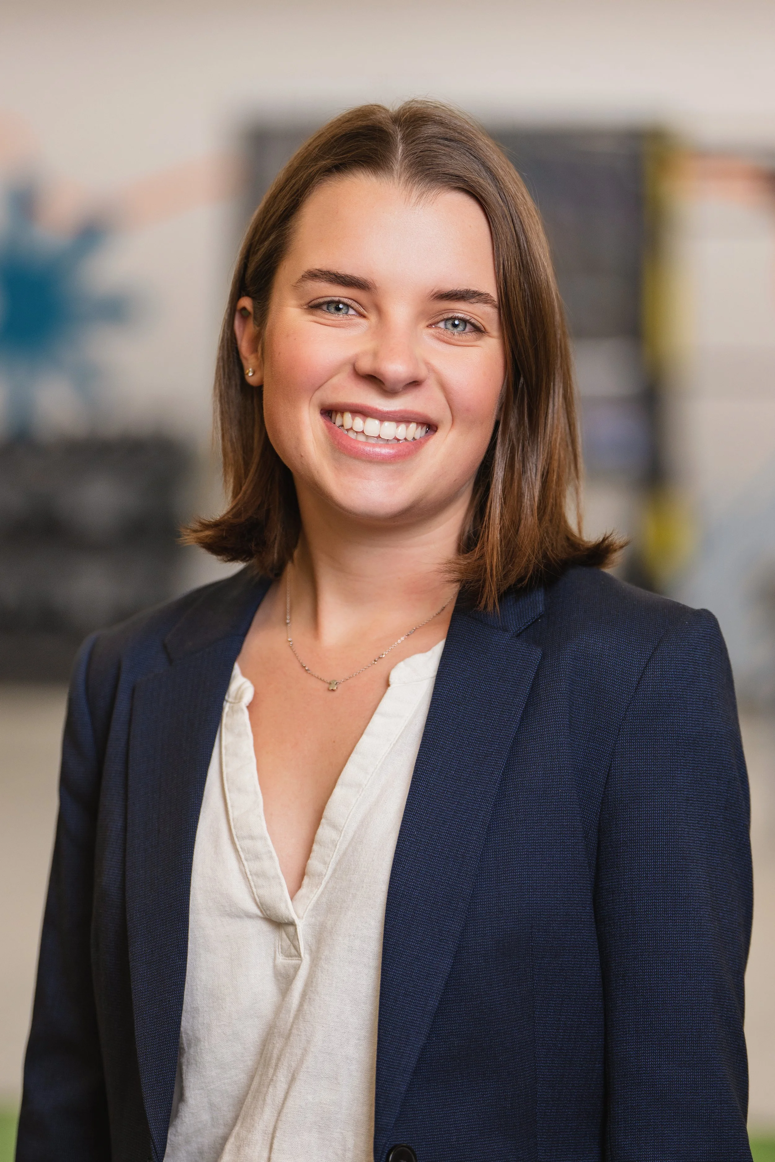 A woman with shoulder-length brown hair, wearing a navy blazer and a cream-colored blouse, smiling in an indoor setting.