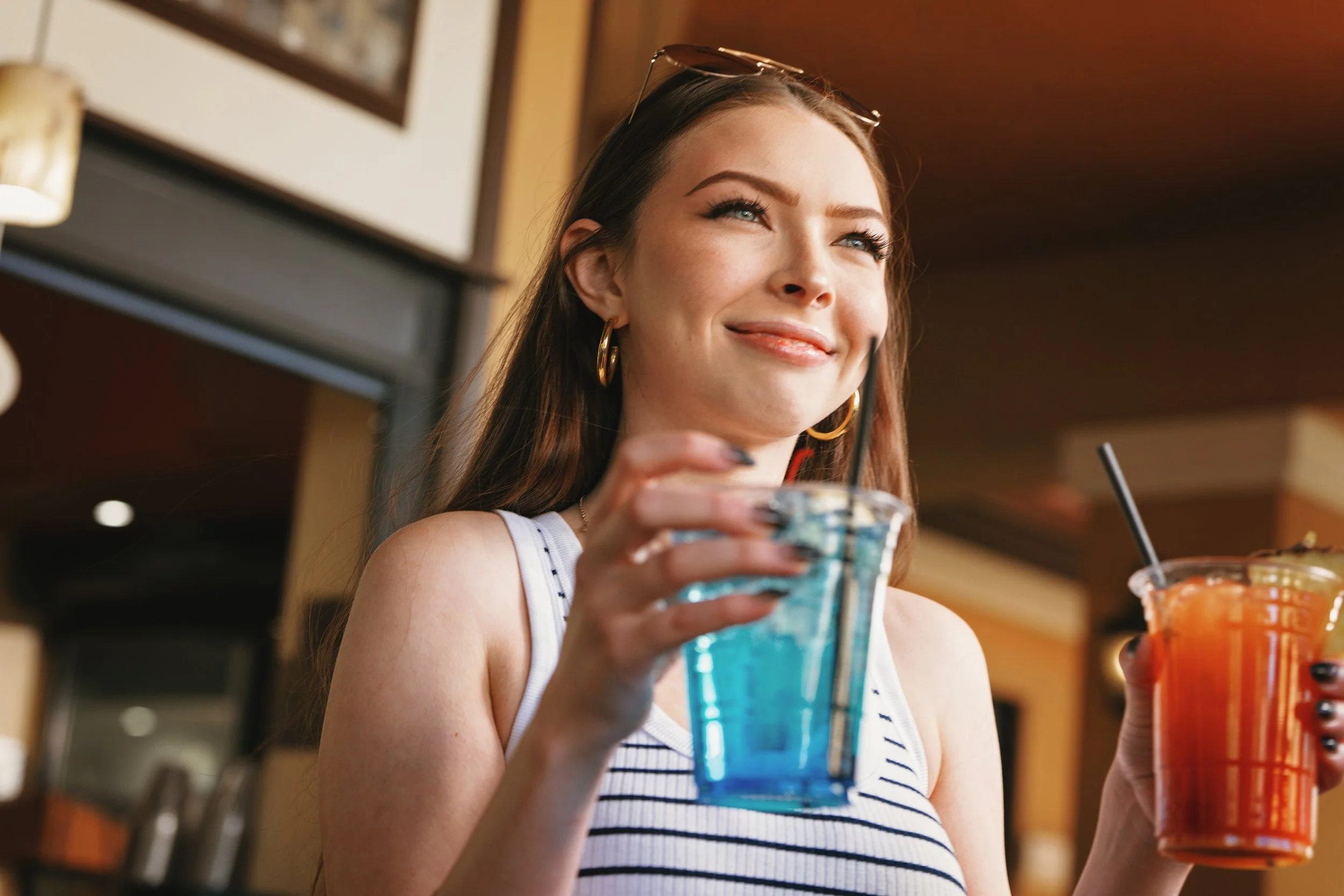 A young woman with long brown hair, wearing a striped tank top and hoop earrings, smiling while holding a blue drink in one hand and a red drink in the other, sitting inside a café or restaurant.