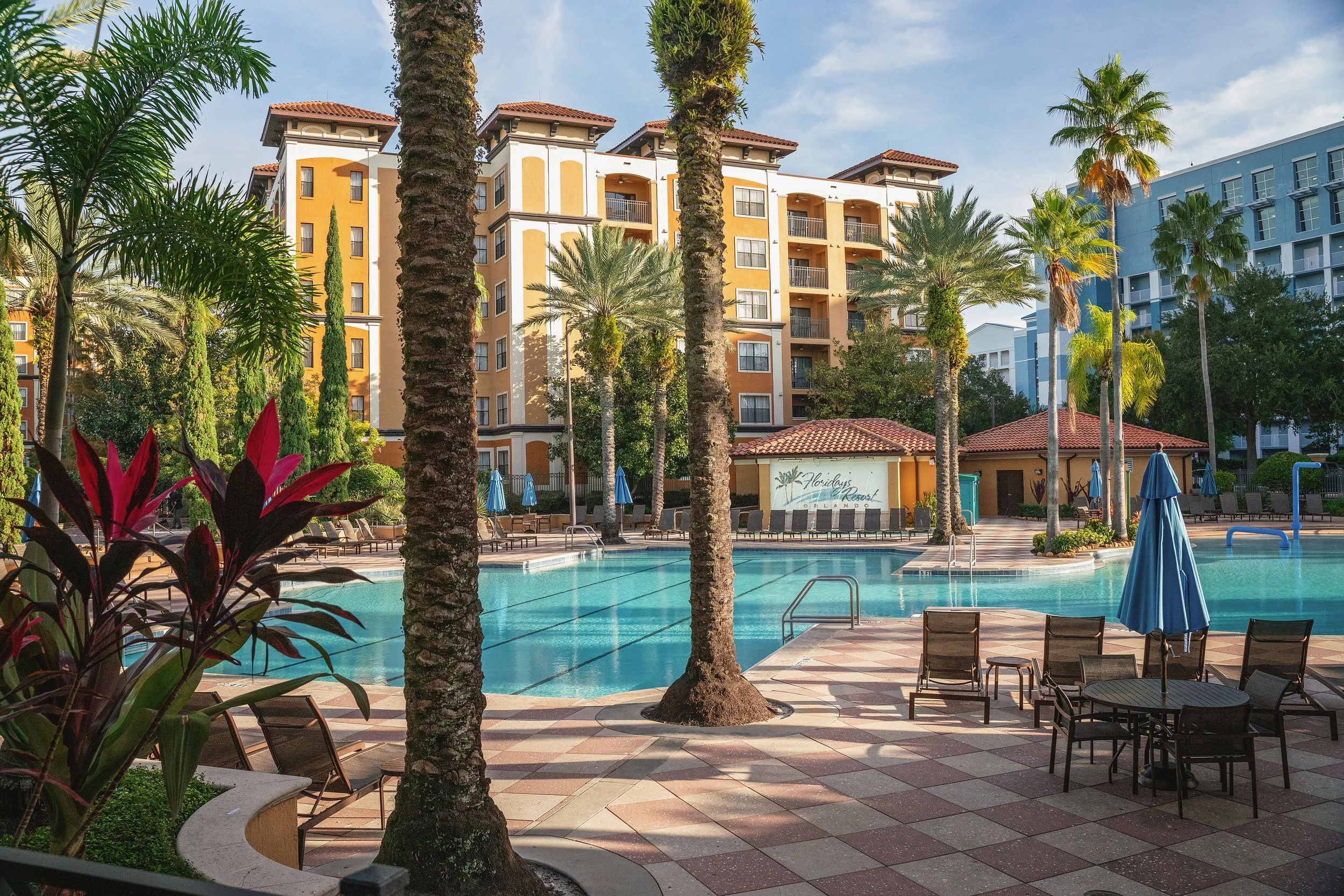 A resort swimming pool area with lounge chairs, umbrellas, and tall palm trees, surrounded by colorful apartment buildings under a blue sky.