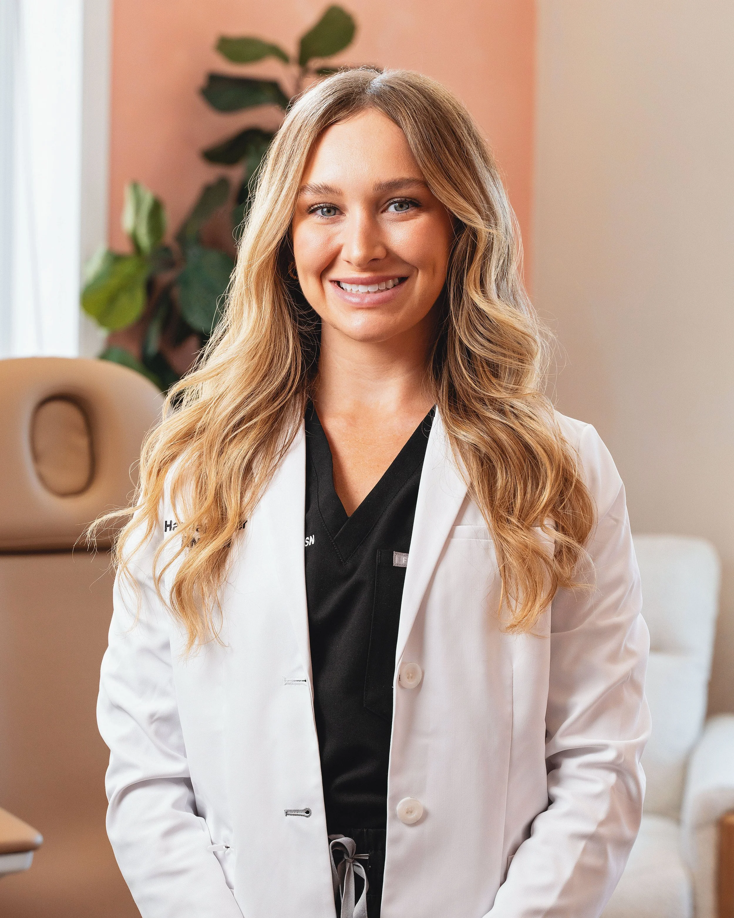 A smiling female healthcare professional wearing a white coat and black scrubs standing in a medical office.