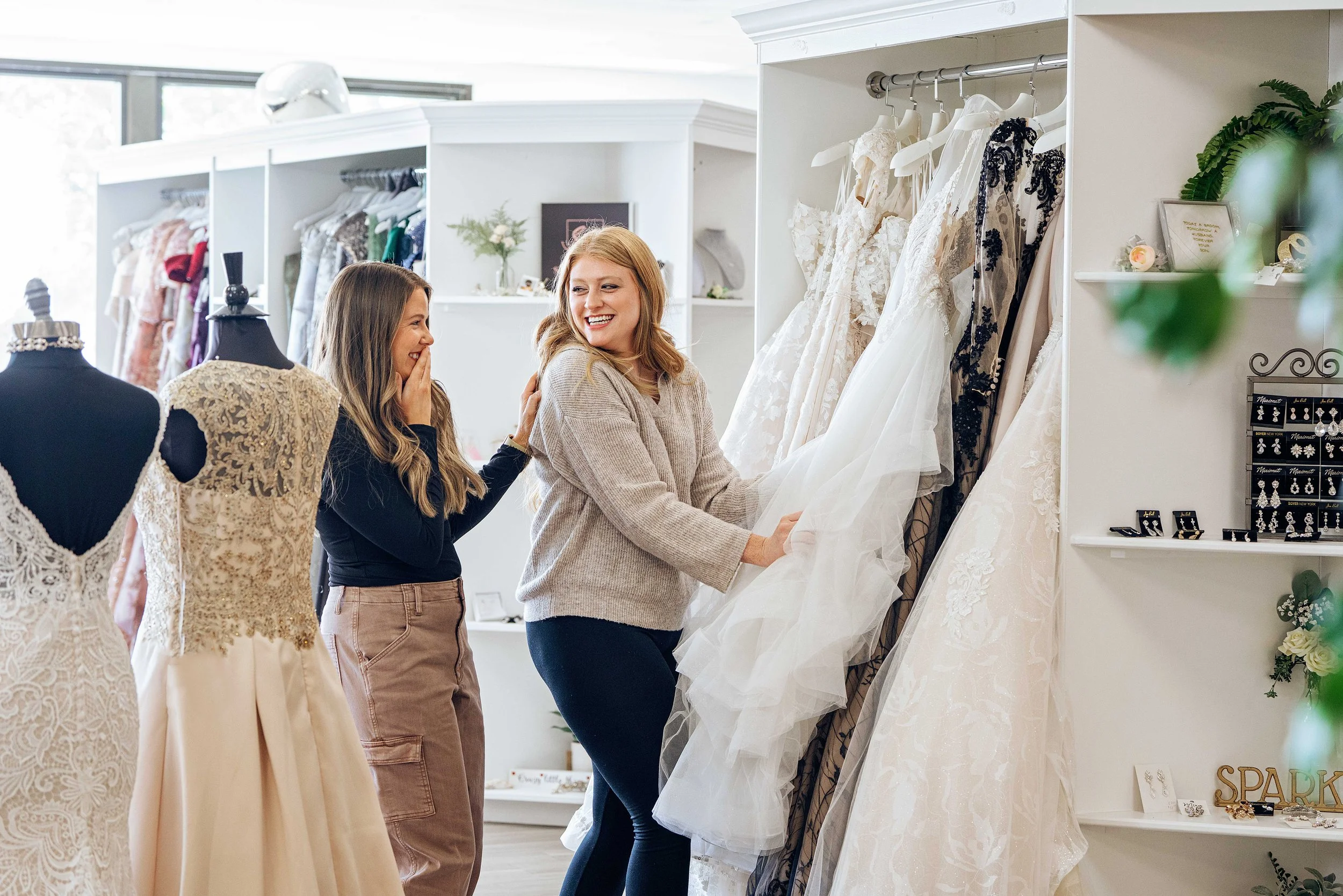 Two women shopping for wedding dresses inside a bridal boutique. One woman, with red hair, is trying on a wedding gown while the other woman, with long brown hair, is smiling and touching the bride's shoulder.