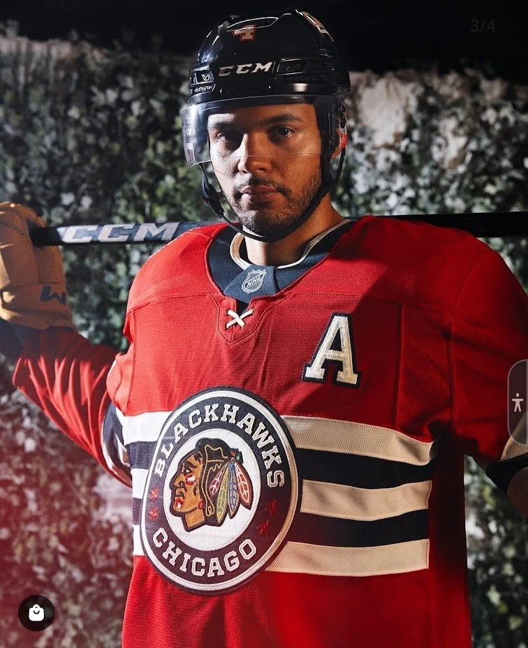 A man wearing a red hockey jersey with the Blackhawks logo, black hockey helmet, and holding a hockey stick over his shoulder.
