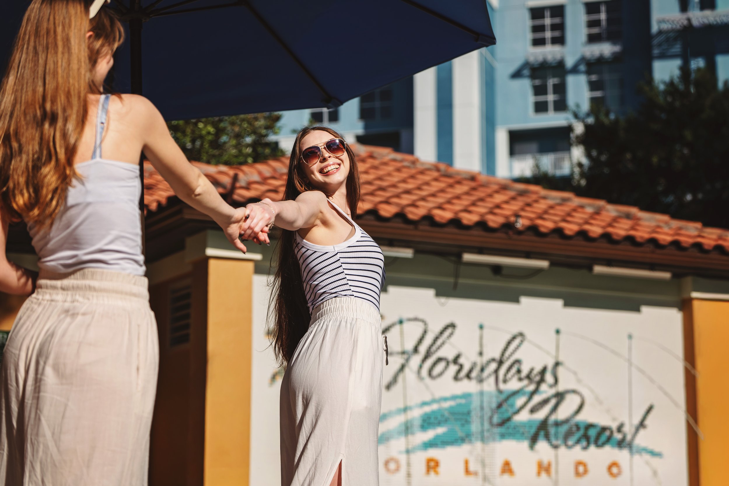 Two women holding hands, smiling, and enjoying a sunny day outside at Florida Days Resort in Orlando. One woman is wearing a striped top and sunglasses, the other is partially visible wearing a white top.