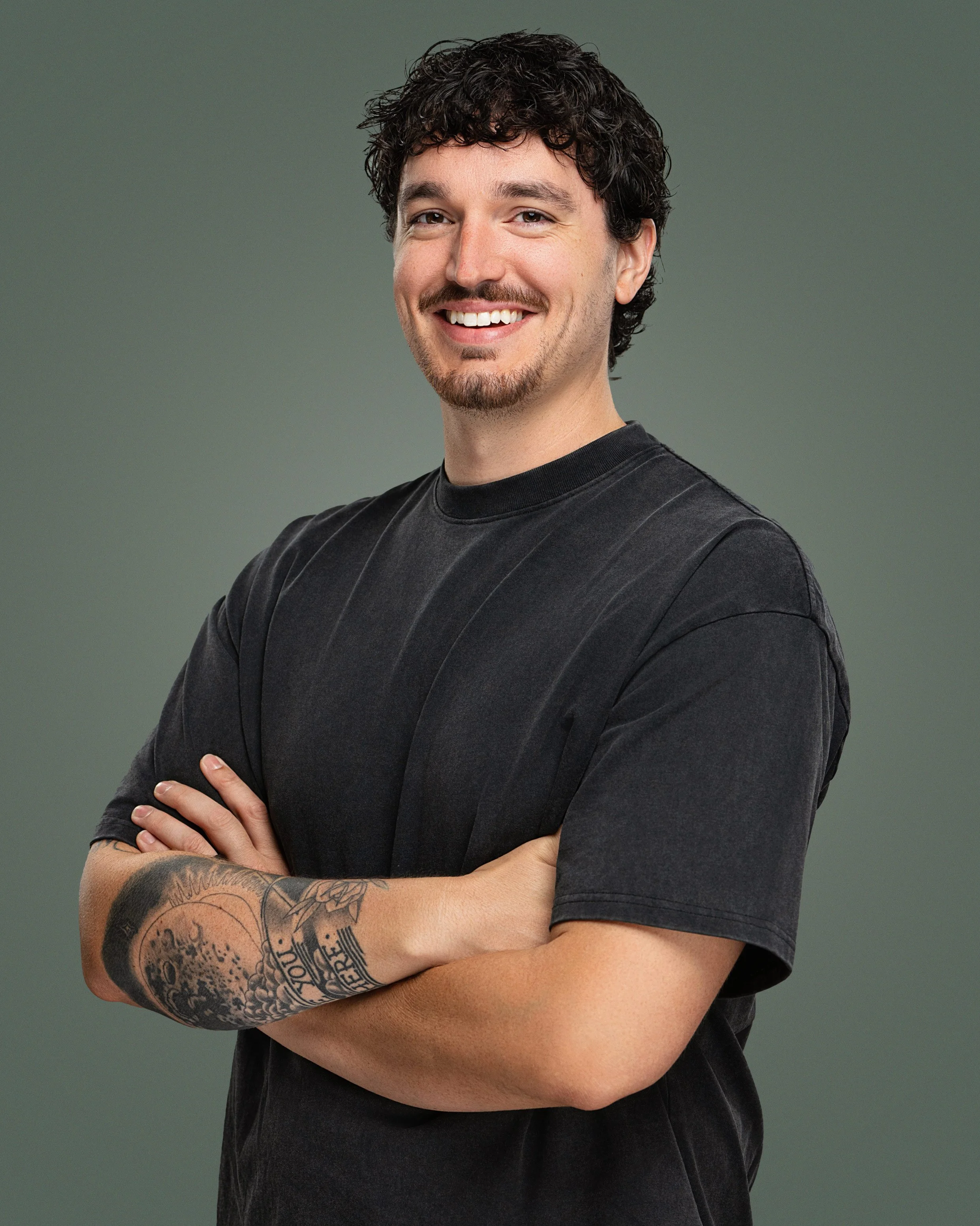 Young man with dark curly hair, light skin, smiling, wearing a black t-shirt, with tattoos on his right arm, standing with arms crossed, against a gray background.