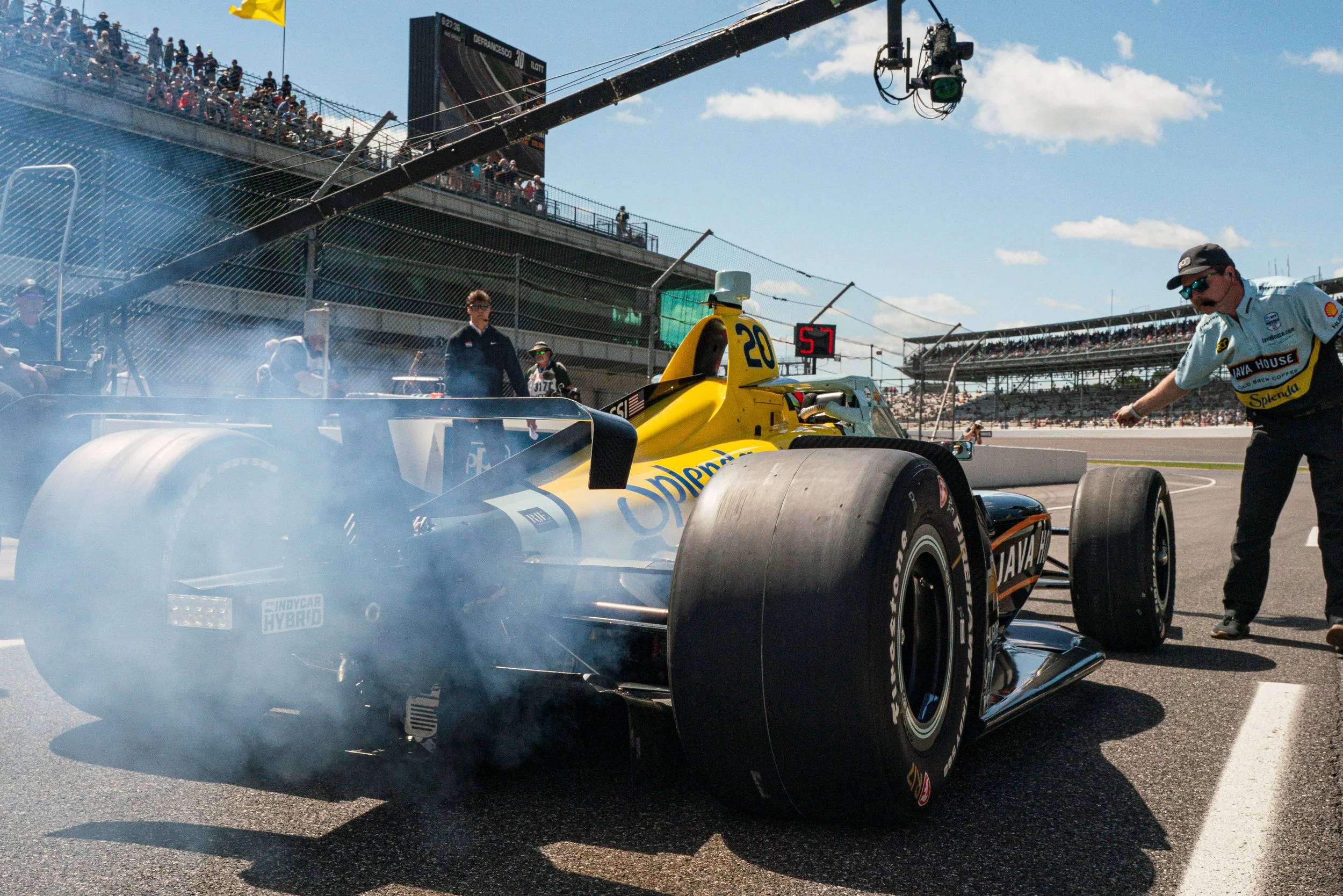 A yellow IndyCar race car on the track during a race, with smoke coming from the tires, and team members standing nearby under a clear sky.