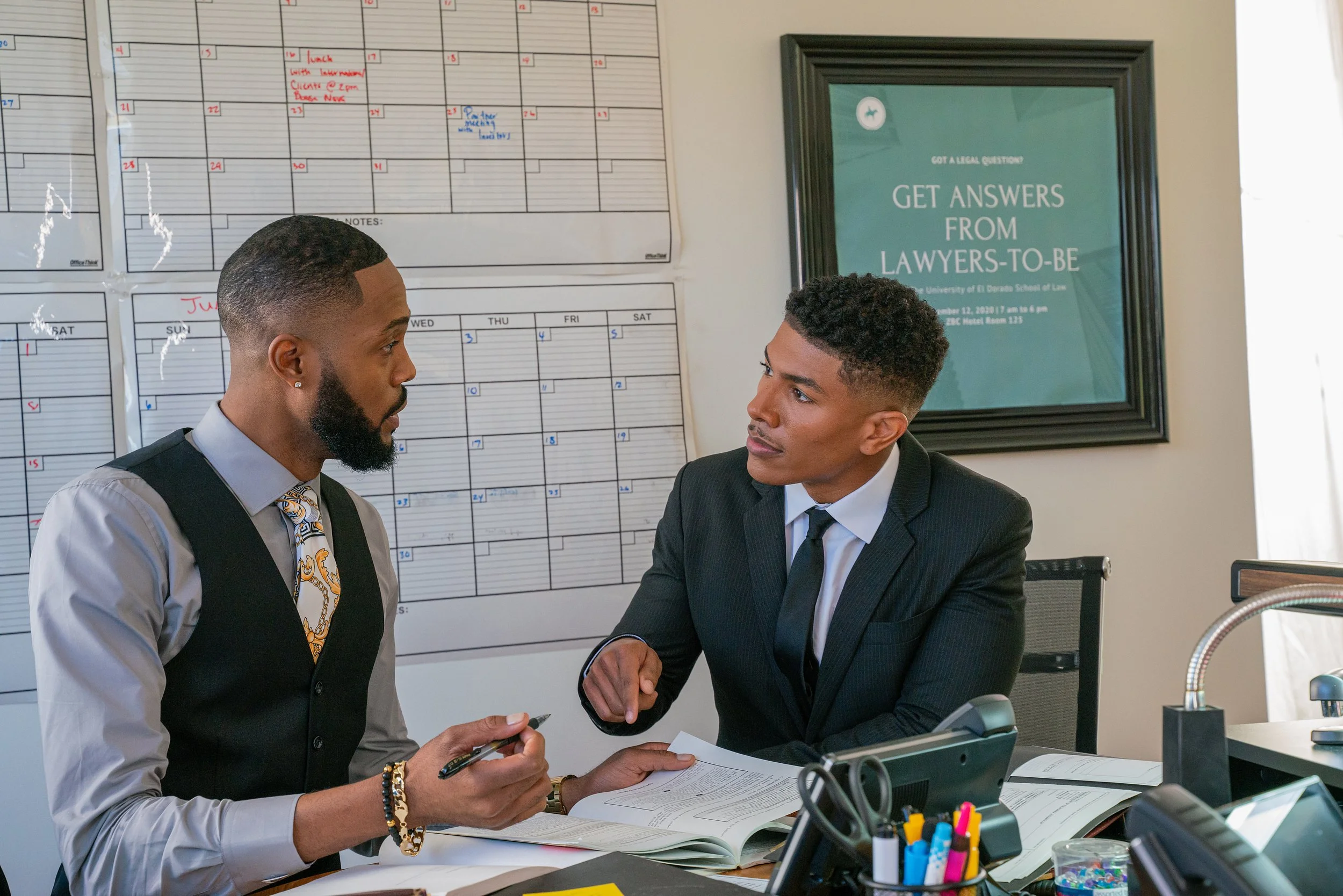 Two men in business suits having a serious conversation in an office. They are seated at a desk with notebooks, papers, and office supplies. In the background, a large wall calendar and a framed sign that reads, 'Get answers from lawyers-to-be' are v