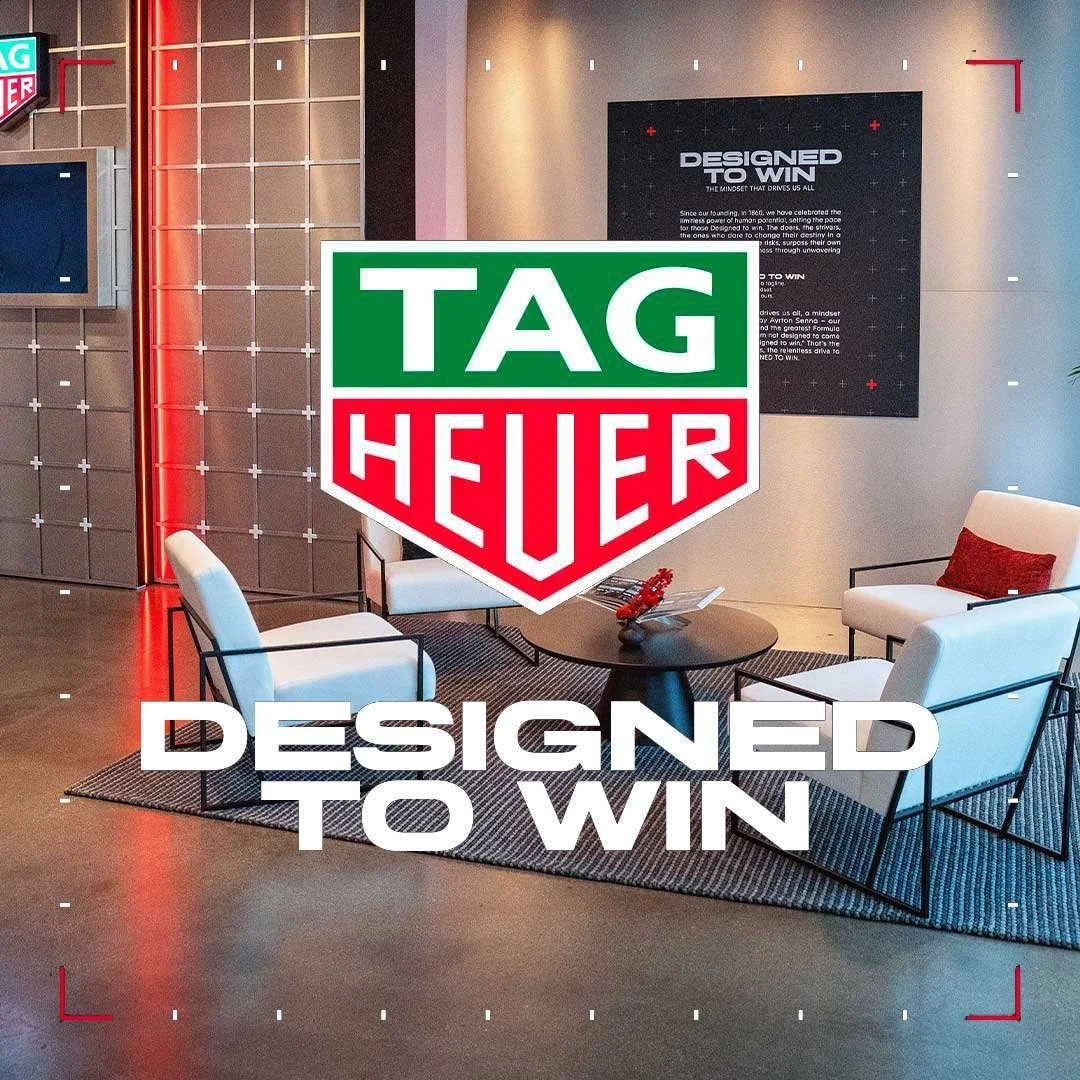 Modern office lounge with white armchairs, a black round coffee table, and a rug. TAG Heuer logo superimposed in front, with a black wall sign titled "Designed to Win" in the background.