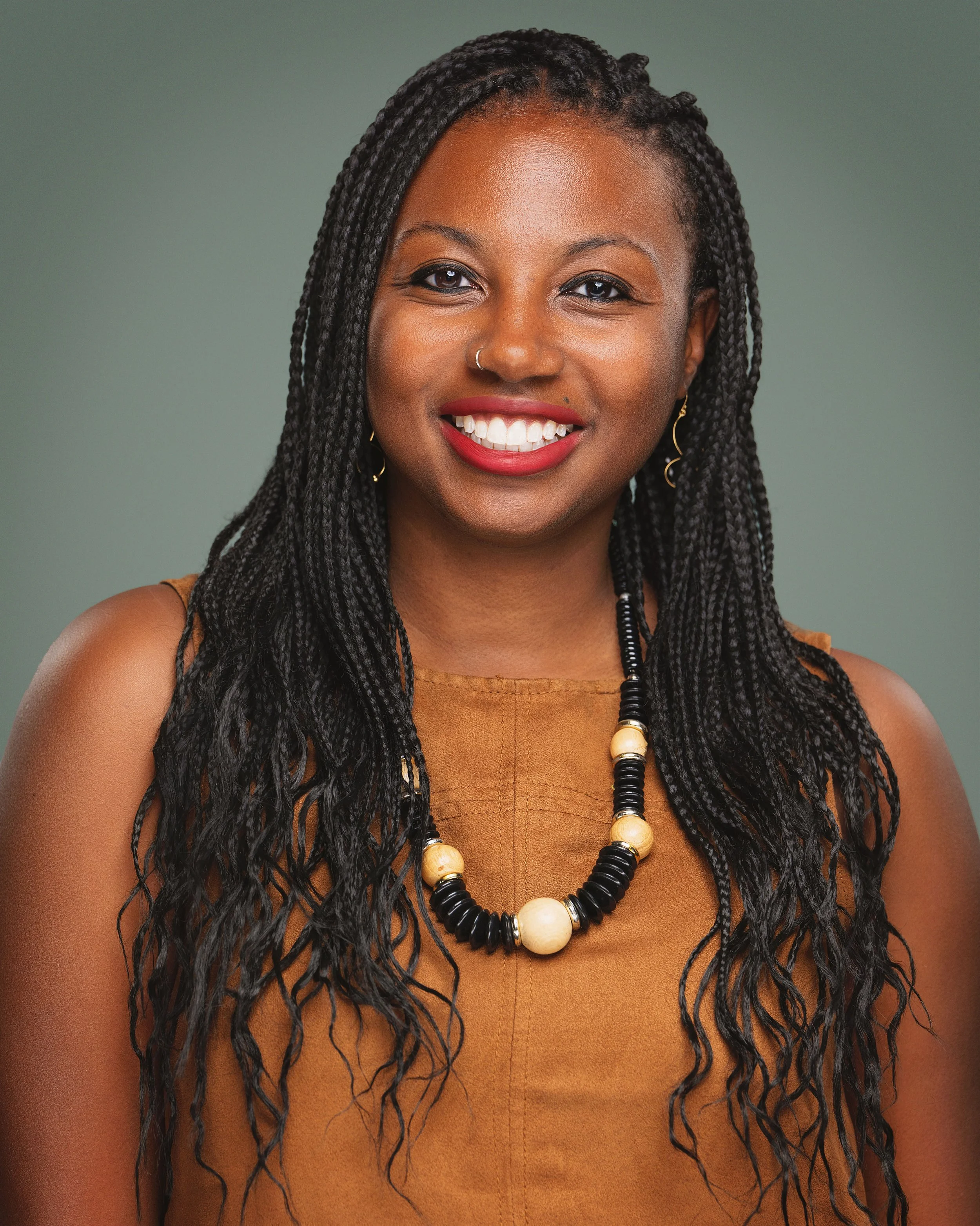 A smiling woman with long, twist braids, wearing a gold-colored top and a black and cream beaded necklace.