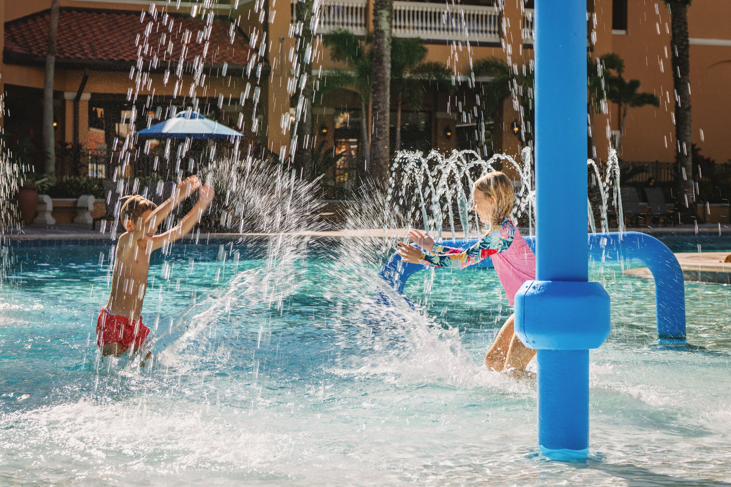 Two children playing in a swimming pool with water jets, splashing water at each other during a sunny day with buildings and trees in the background.