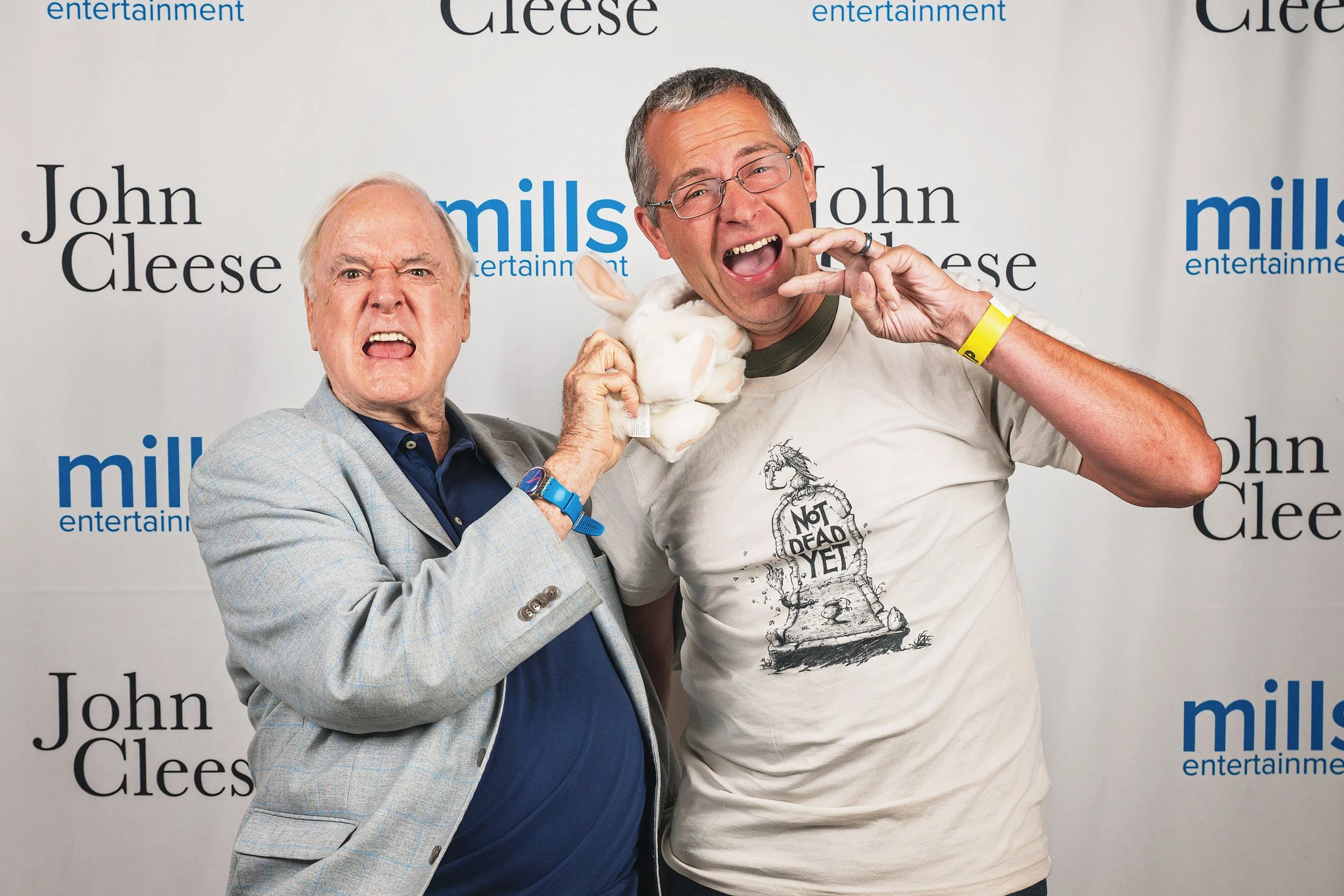 Two men at a comedy event, John Cleese is older with white hair in a plaid blazer, making an angry face, and the other is younger with glasses in a T-shirt, smiling and holding a stuffed rabbit.