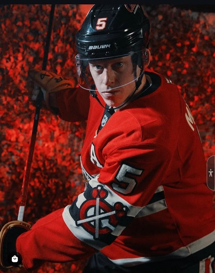 A male hockey player in full gear, wearing a red jersey with the number 5, and a black helmet, skating on ice with a red background.