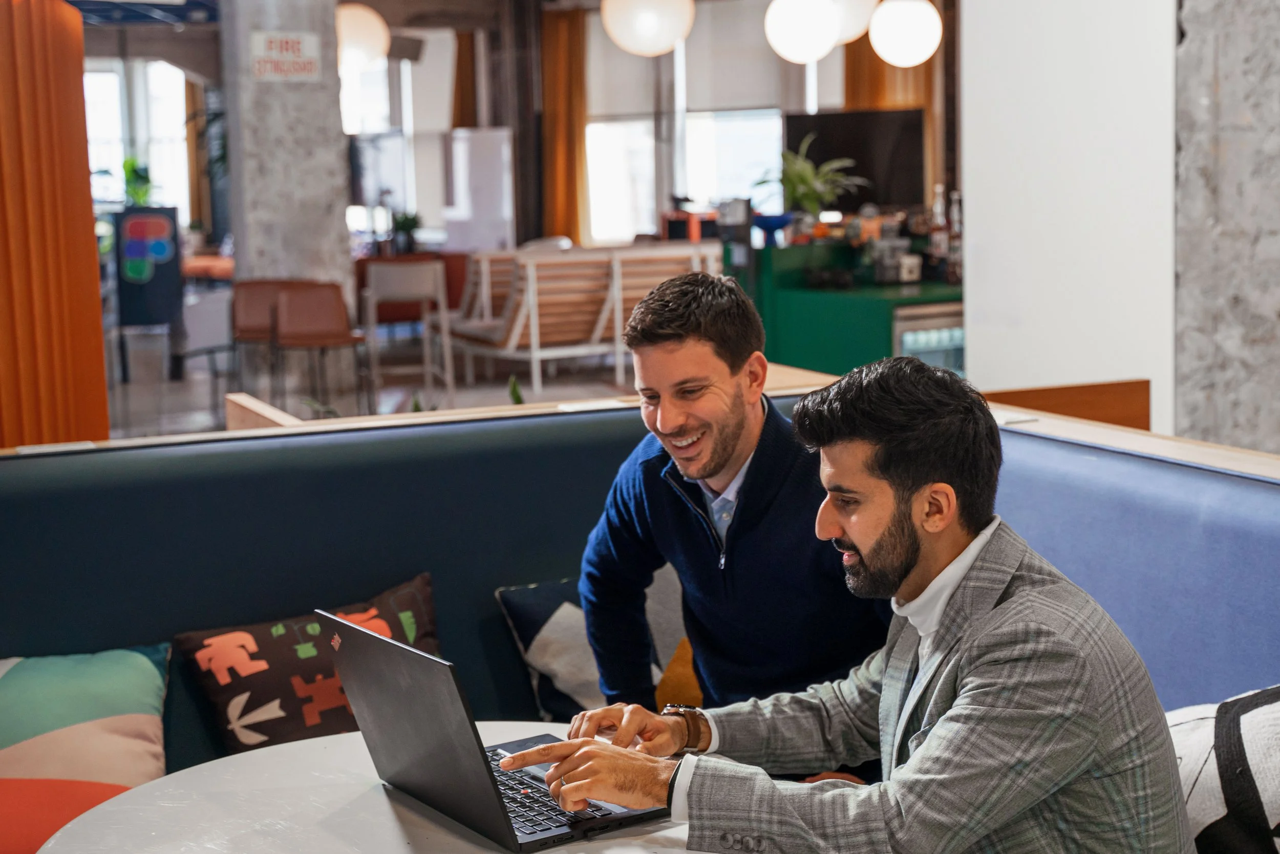 Two men in business attire working on a laptop in a modern, cozy office or cafe setting, with seating and large windows in the background.
