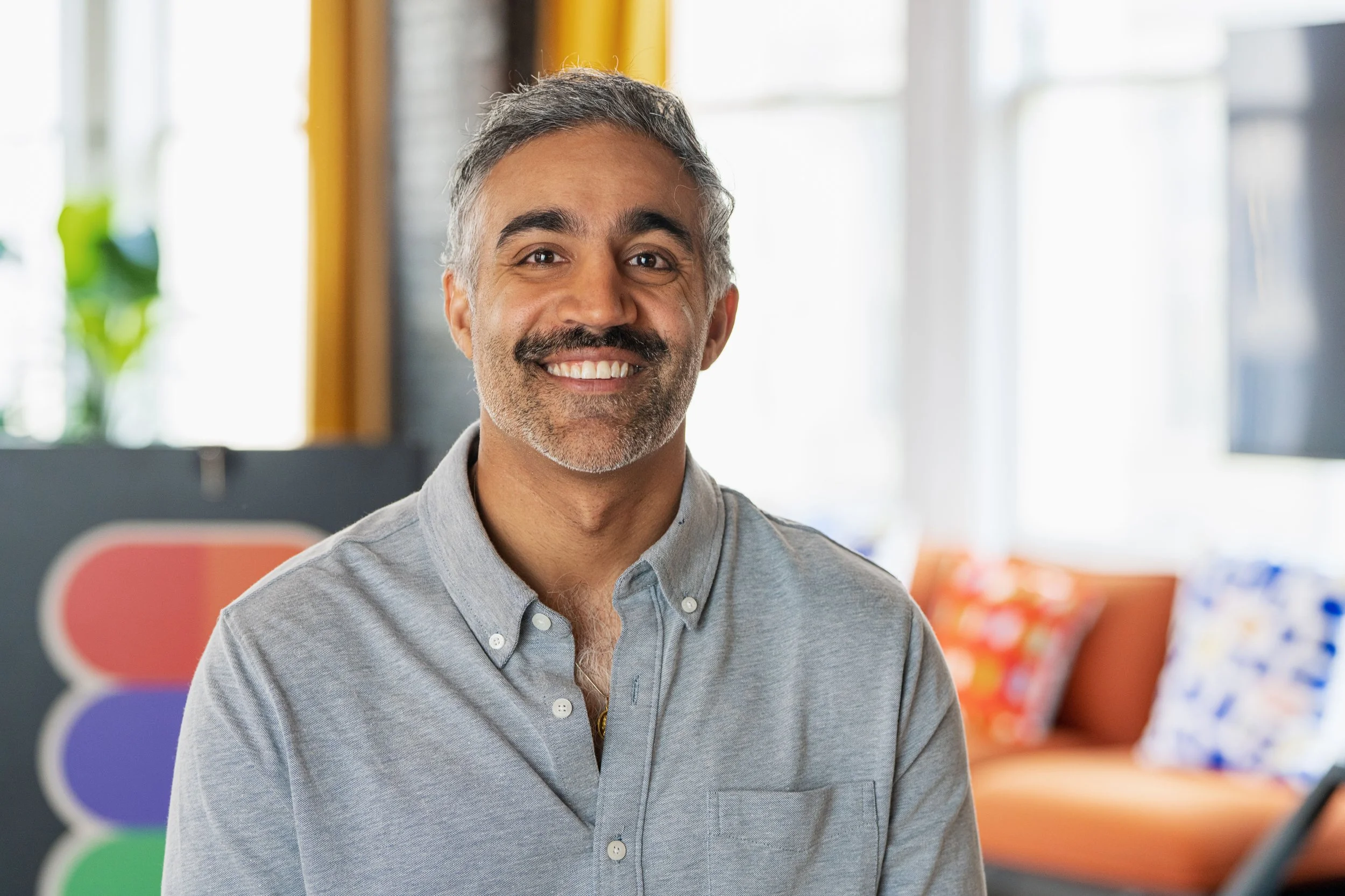 A smiling middle-aged man with salt and pepper hair and a beard, wearing a light gray button-up shirt, sitting in a colorful and bright office space.