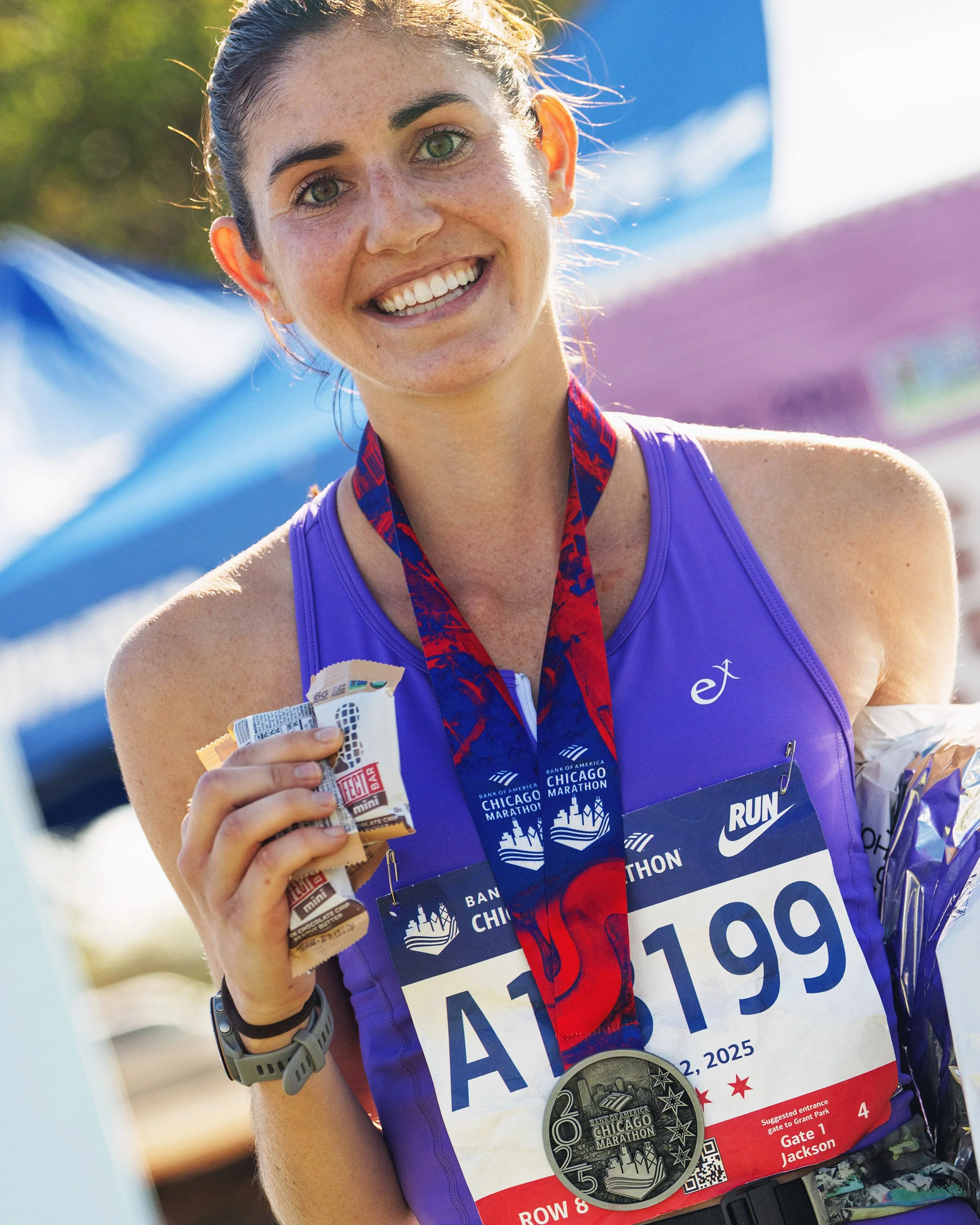 A smiling woman wearing a blue athletic tank top with a marathon bib, medal, and snacks, celebrating her finish at the Chicago Marathon.
