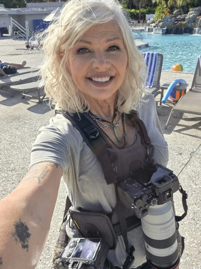 Woman with blonde, curly hair smiling, taking a selfie near a pool, wearing photography gear including a camera with a large lens, a second camera, and sitting by poolside chairs.