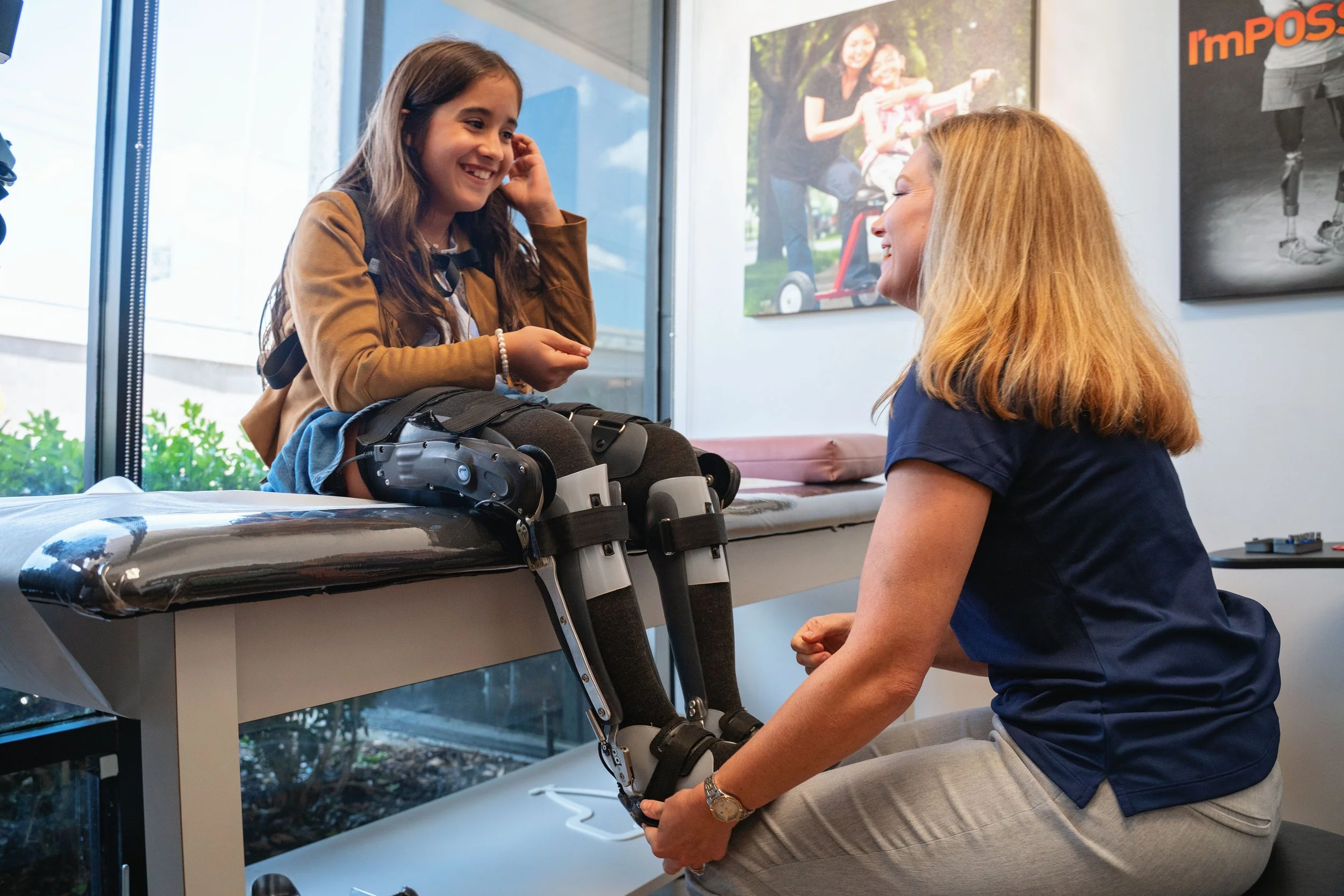 A young girl in a wheelchair talking with a woman, possibly a physical therapist, in a medical office. The girl is smiling and wearing braces on her legs; the woman is kneeling, holding the girl's leg. There are posters on the wall.