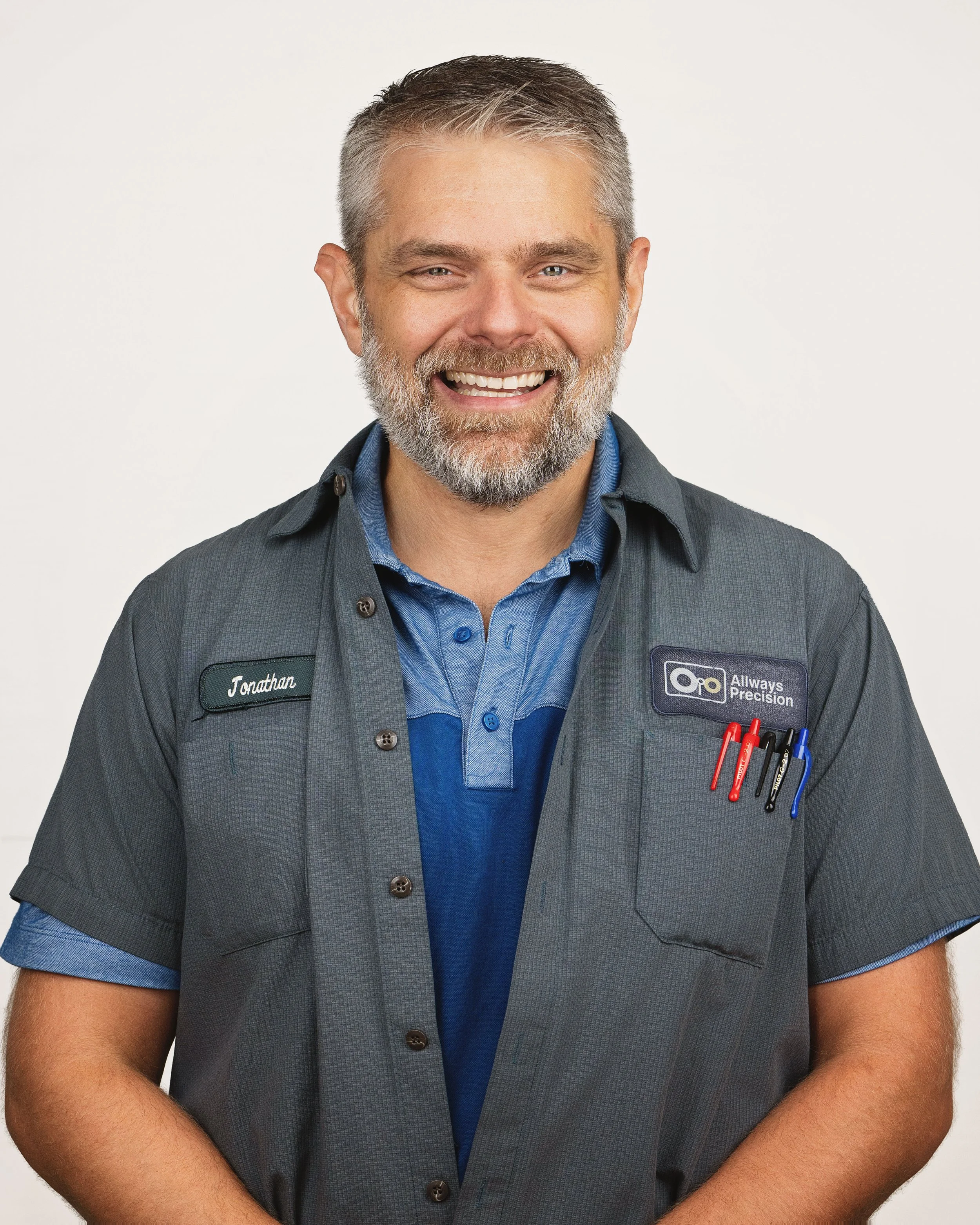 A smiling man with gray hair and a beard wearing a gray uniform shirt with name tag 'Jonathan' and a company logo 'Allways Precision', with pens in his shirt pocket.