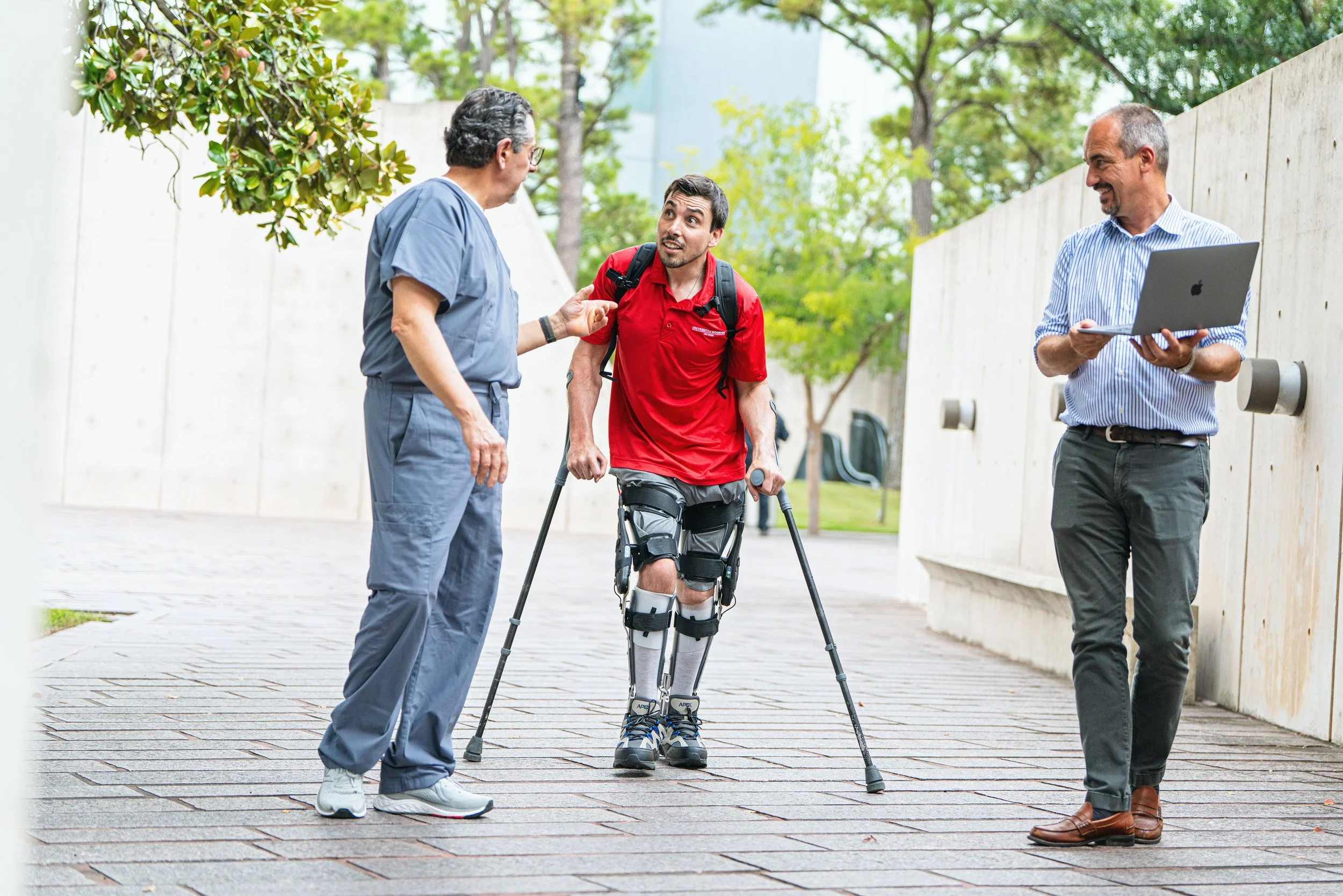 A young man with a prosthetic leg and crutches talking to a doctor outside, while another man with a laptop observes, in a sunny outdoor setting.