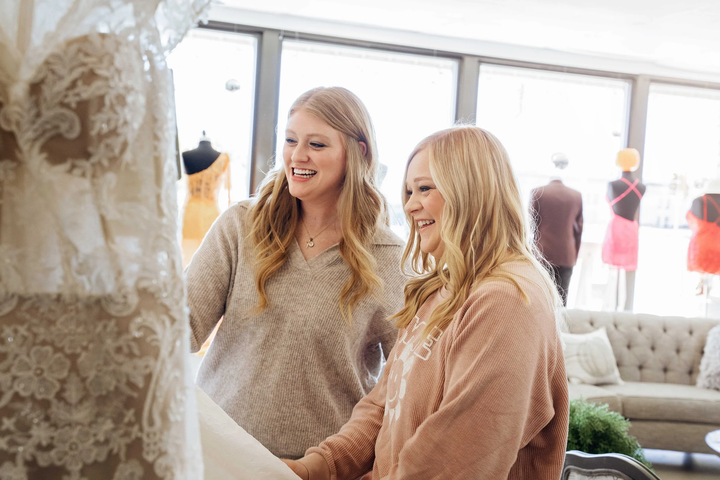 Two women shopping for wedding dresses in a store, smiling and looking at a lace wedding gown.
