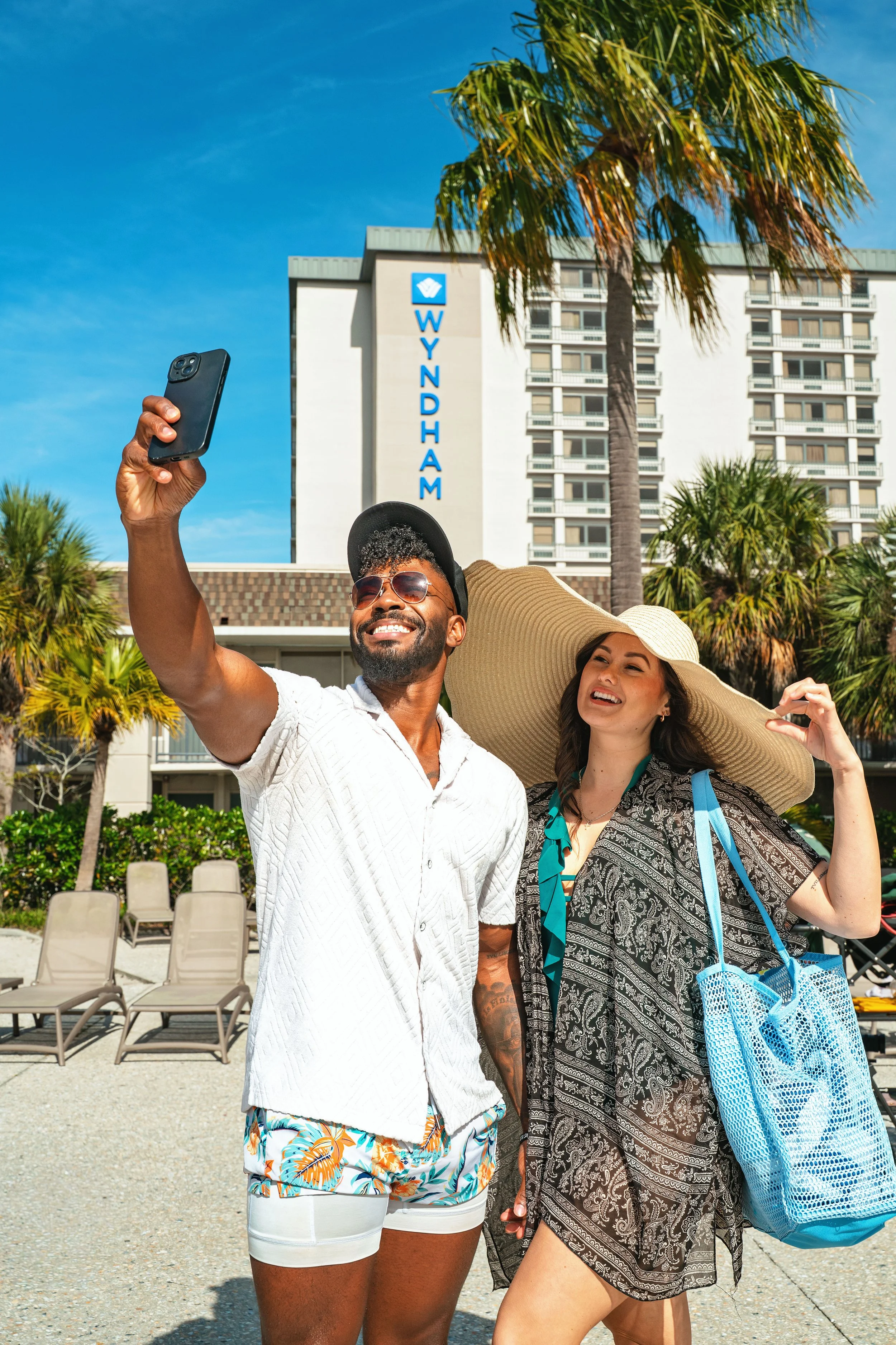 Two people taking a selfie in front of a Wyndham hotel with palm trees and a clear blue sky.