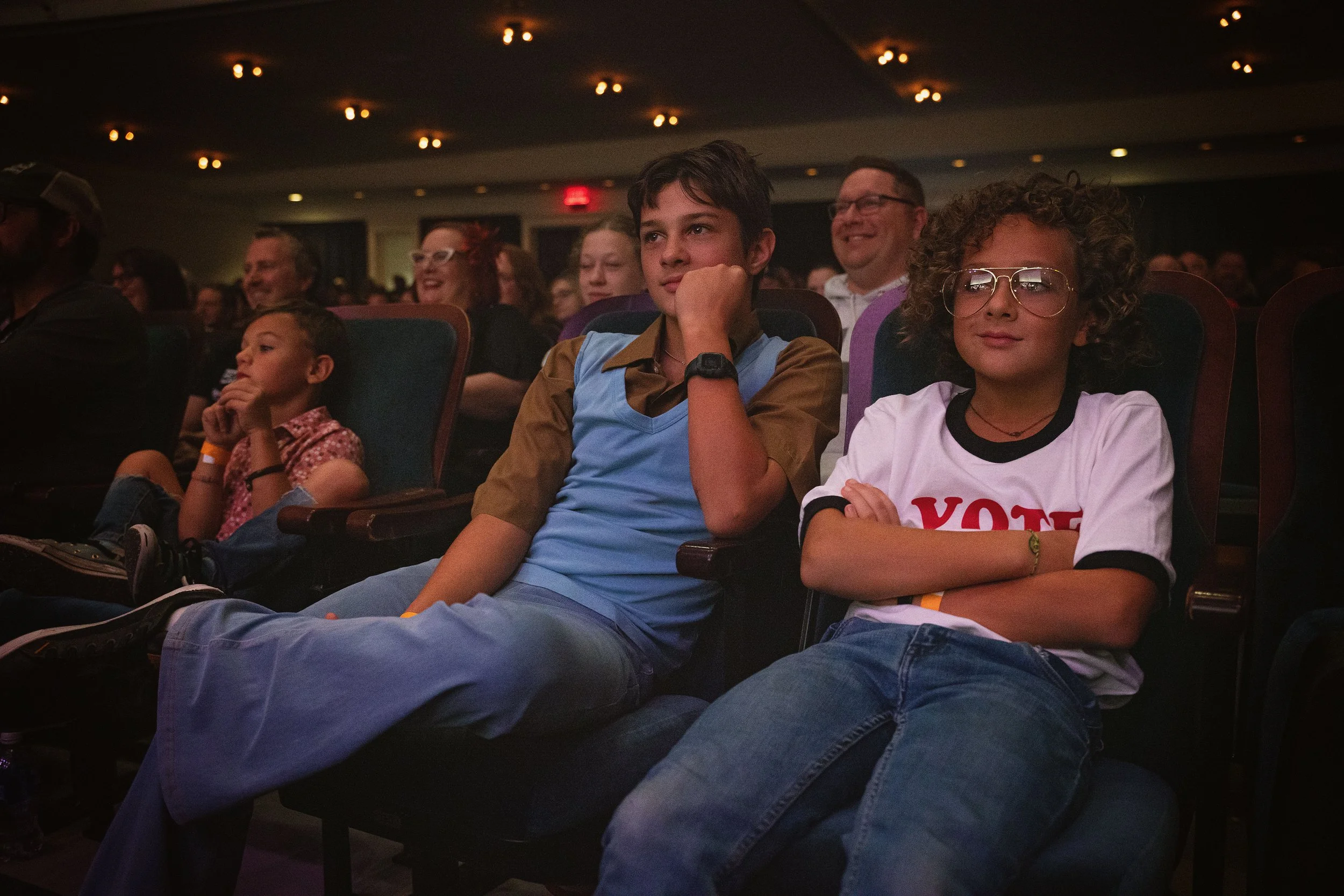 Audience members sitting in theater seats during a presentation or performance, with children and adults looking attentive and engaged.