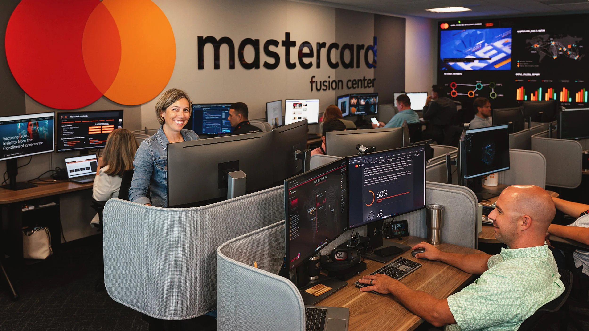 Inside the Mastercard fusion center, with multiple employees working at computers and monitoring screens displaying data, maps, and charts; the Mastercard logo is prominently displayed on the wall.
