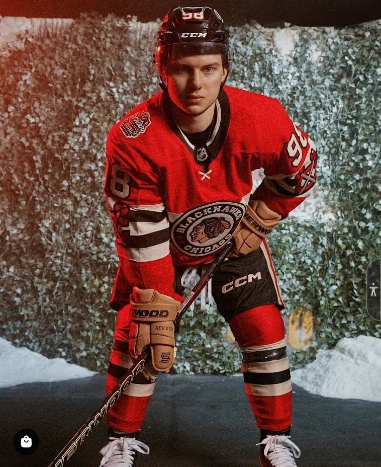 A hockey player in red and black uniform with a black helmet, standing on ice with a background of snow and trees, holding a hockey stick.