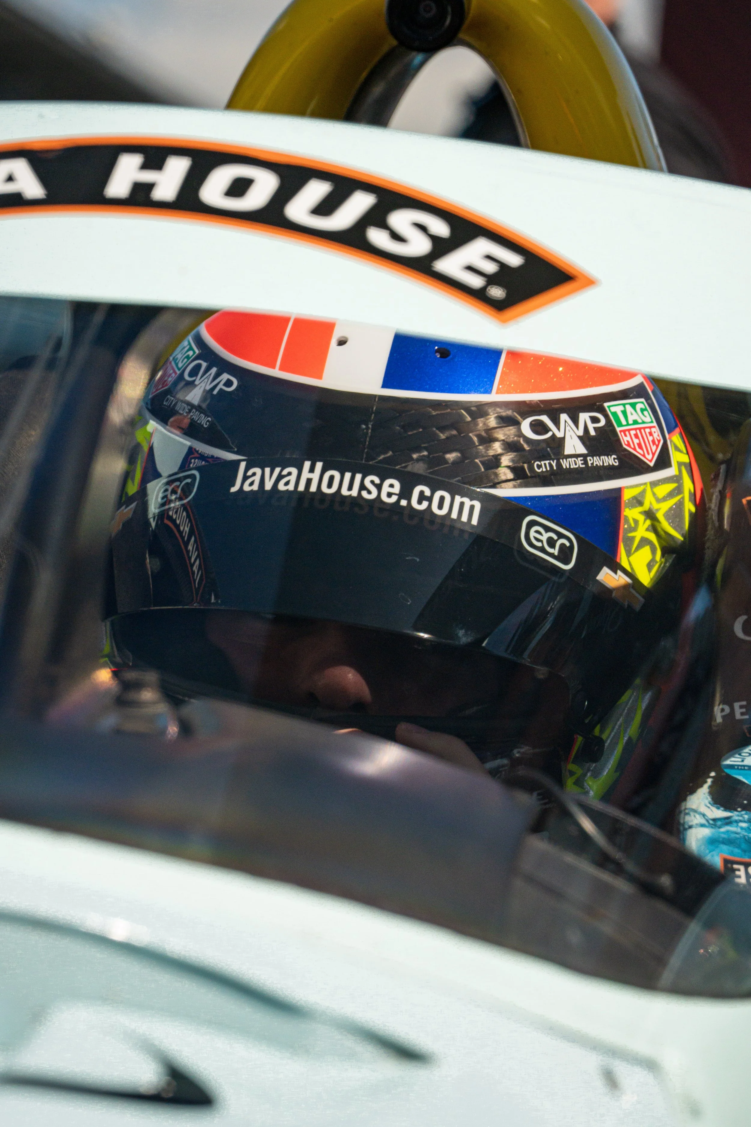Race car driver wearing a helmet with the web address JavaHouse.com, inside a race car with the windshield reflecting the driver’s face.