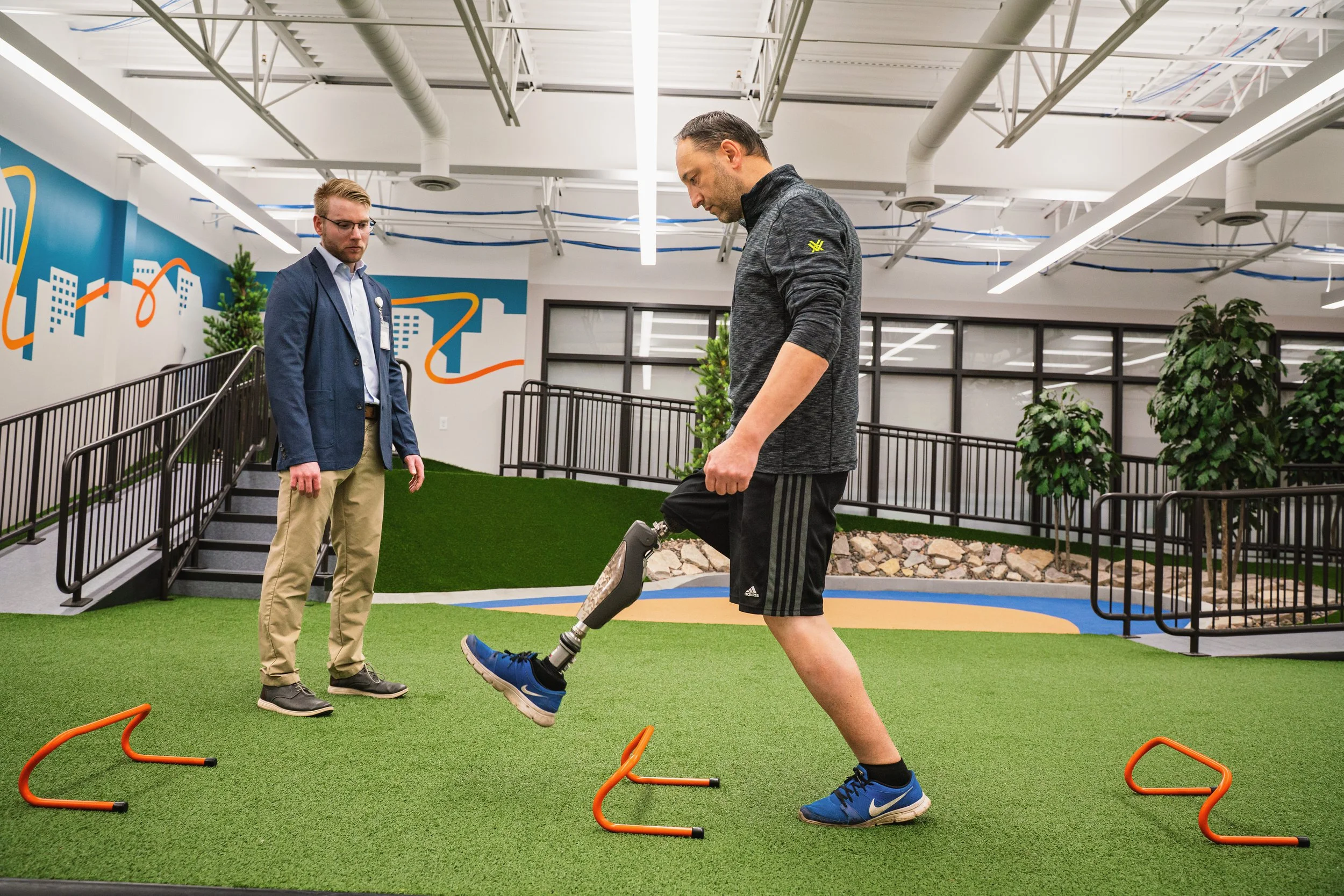 A man with a prosthetic leg participating in a physical therapy or rehabilitation session indoors, with a trainer or therapist observing, in an indoor facility decorated with trees, grass, and colorful wall art.