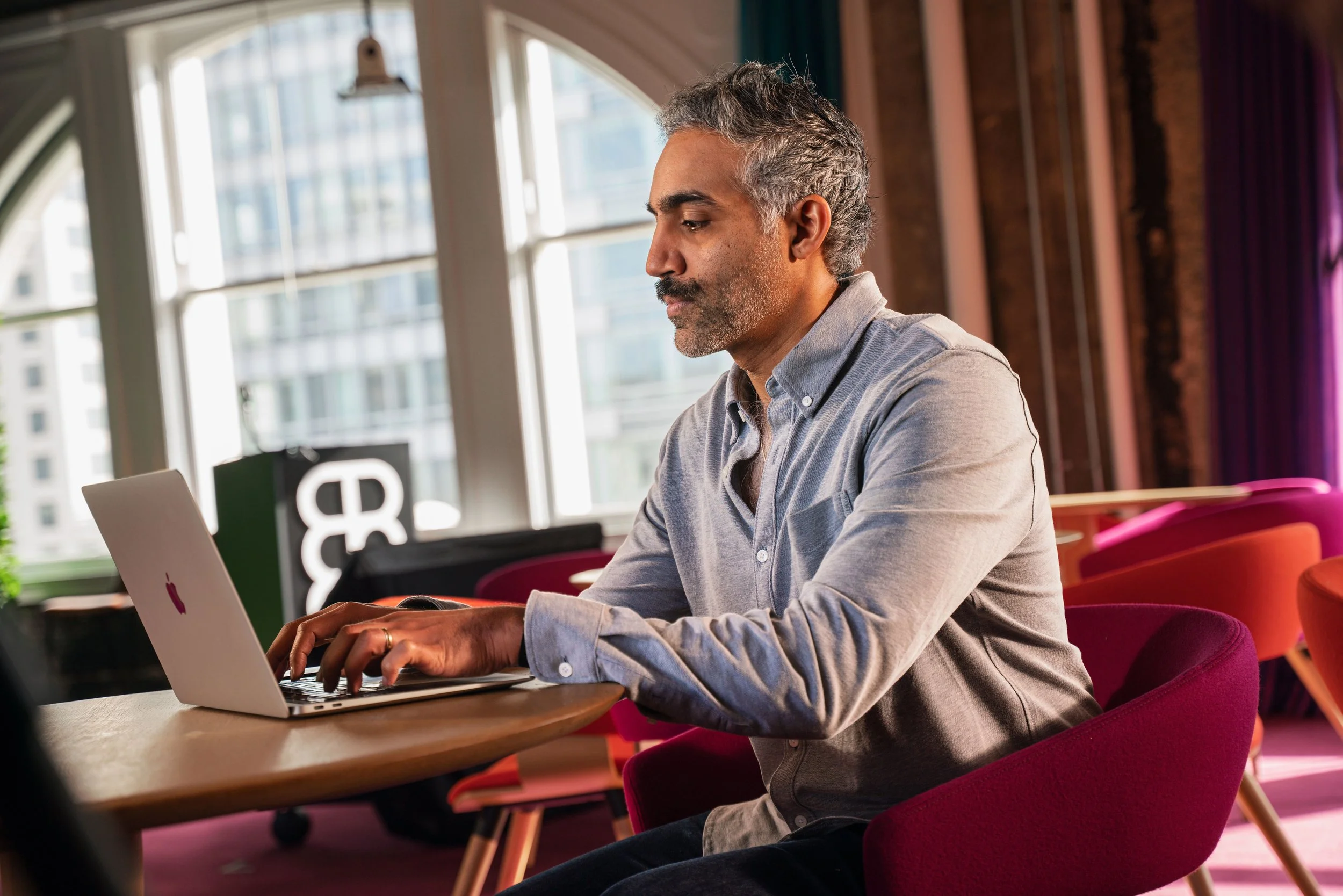 A middle-aged man with gray hair and a beard working on a silver MacBook laptop at a round wooden table in a brightly lit, modern space with large windows and colorful chairs.