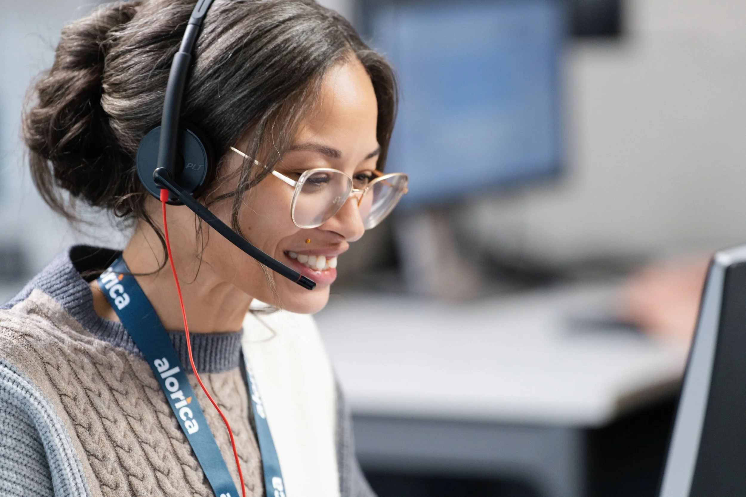 A woman with dark hair tied back, wearing glasses and a headset, smiling while looking at a computer screen. She has a lanyard that says 'alorica'.