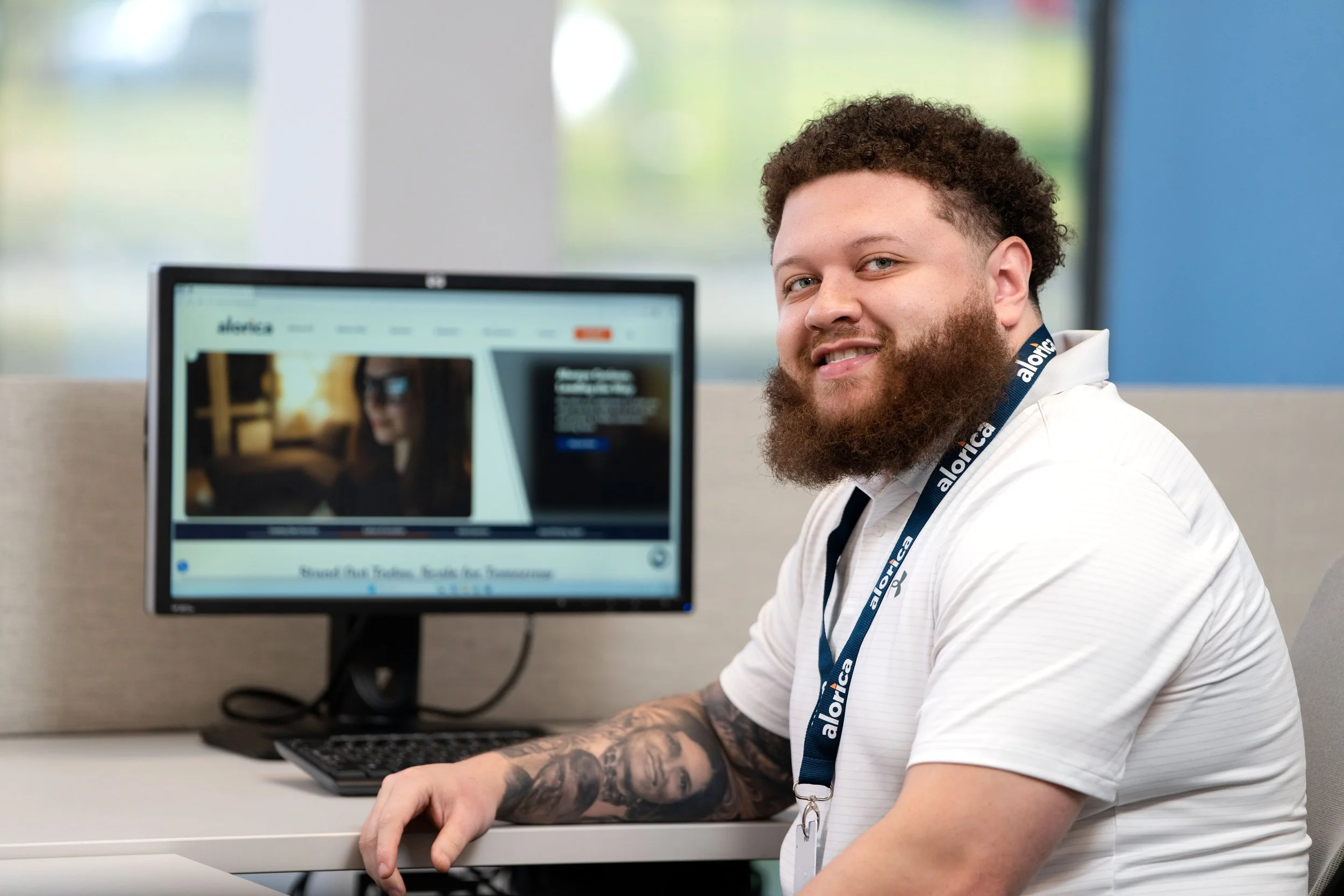 A man with curly hair and a beard sitting at a desk using a computer, wearing a white shirt and a lanyard that says 'alorica'.