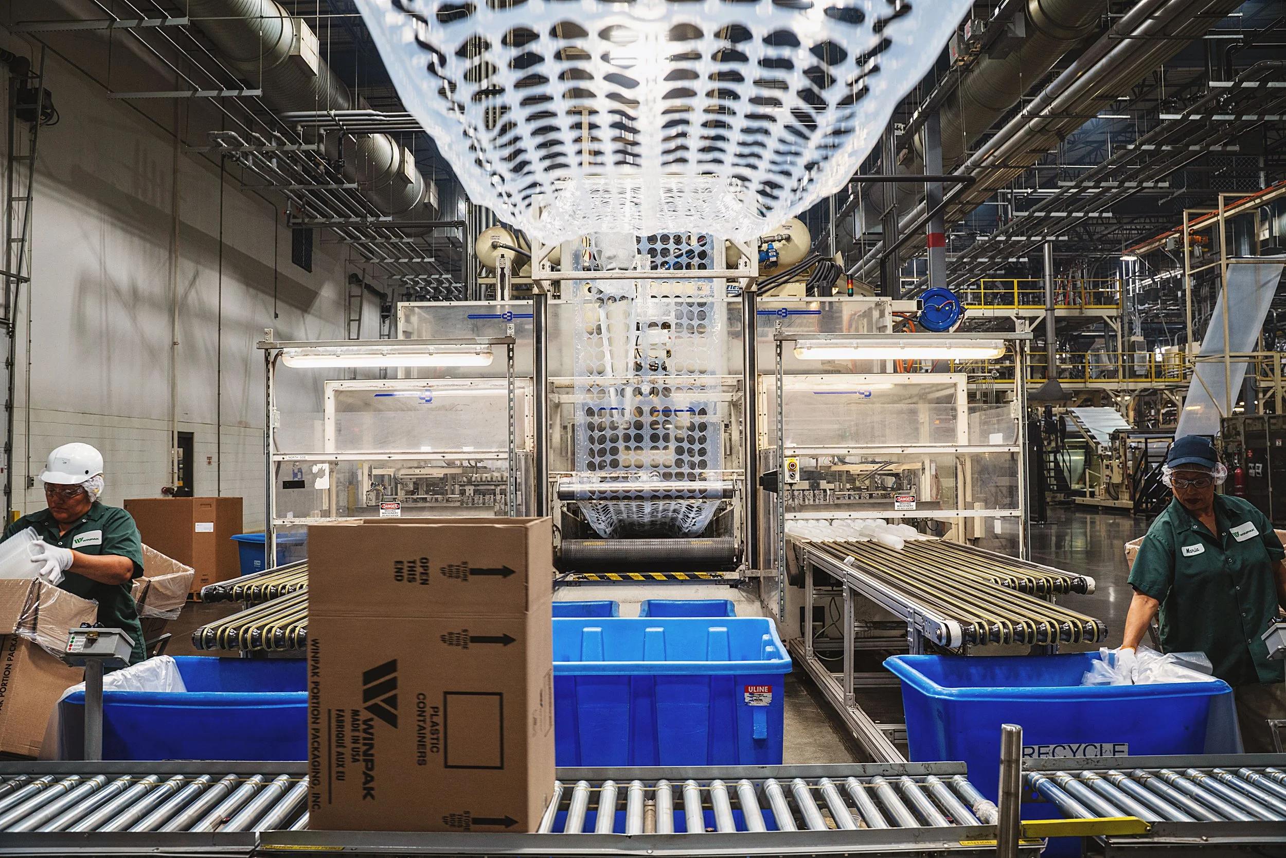 A manufacturing or processing facility with conveyor belts, workers wearing safety gear, and packaging materials, including cardboard boxes and recycling containers.