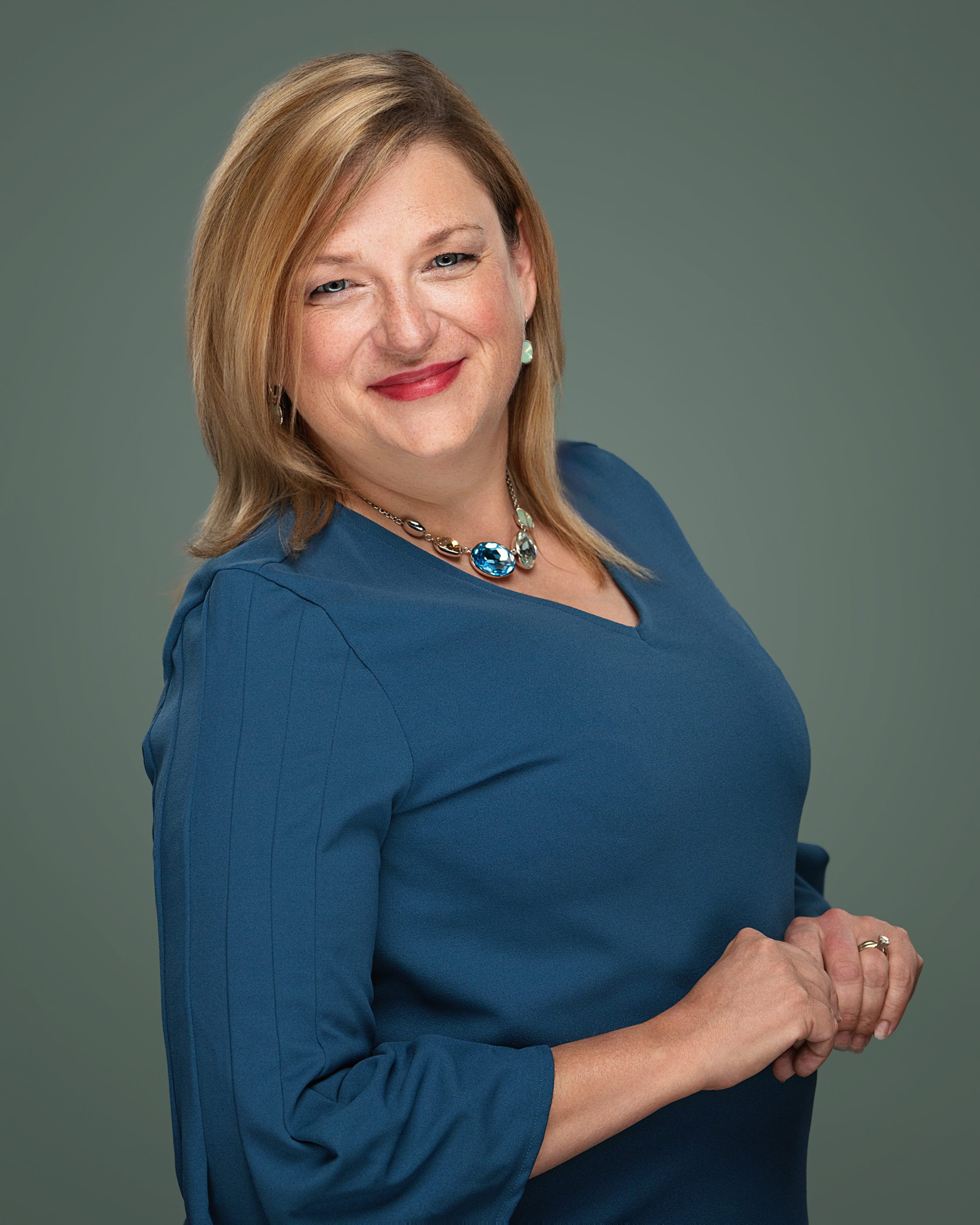 A woman with blonde hair, wearing a blue dress and jewelry, smiling at the camera against a gray background.