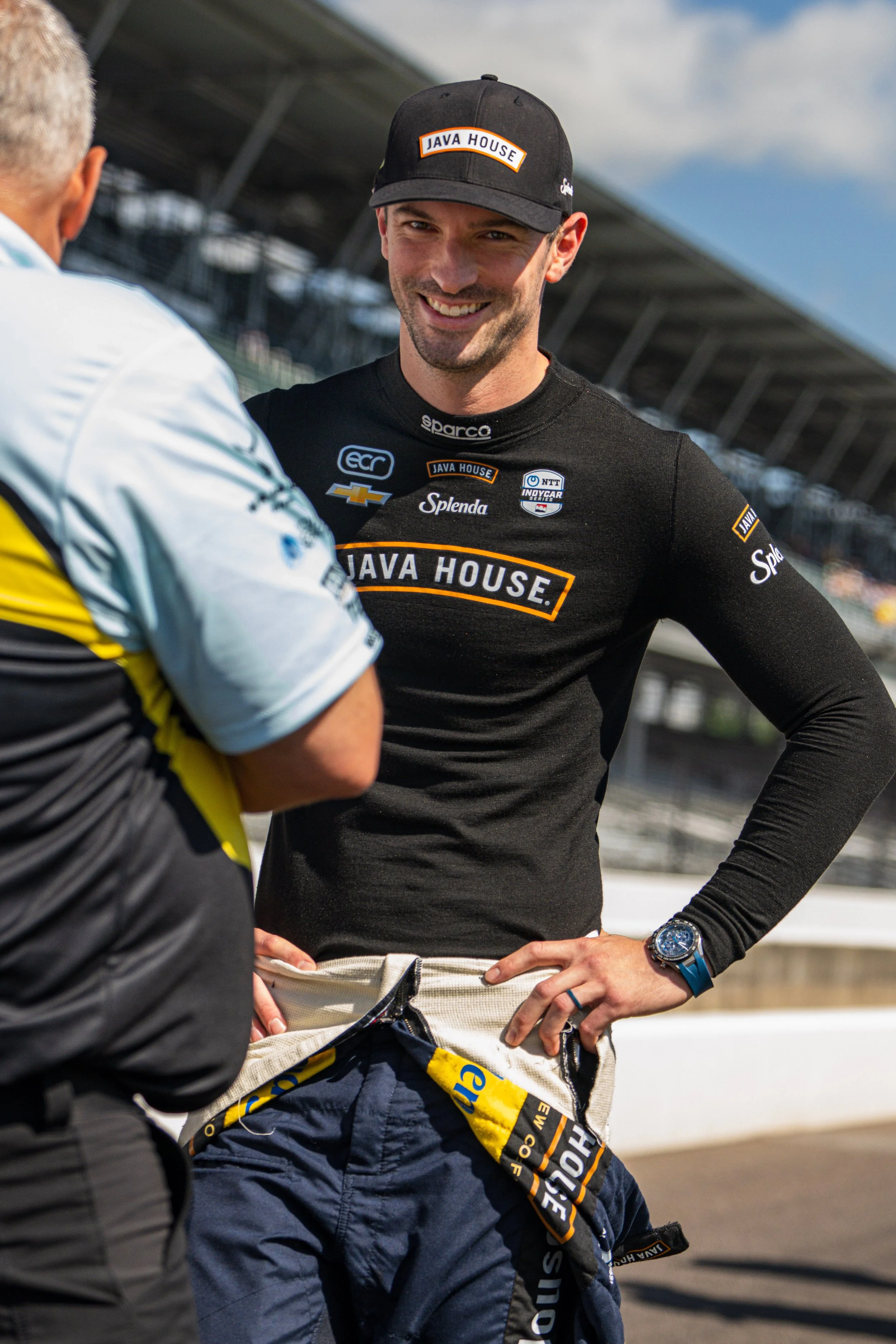 A race car driver in a black Java House shirt and cap smiling and talking to a team member at the racetrack.