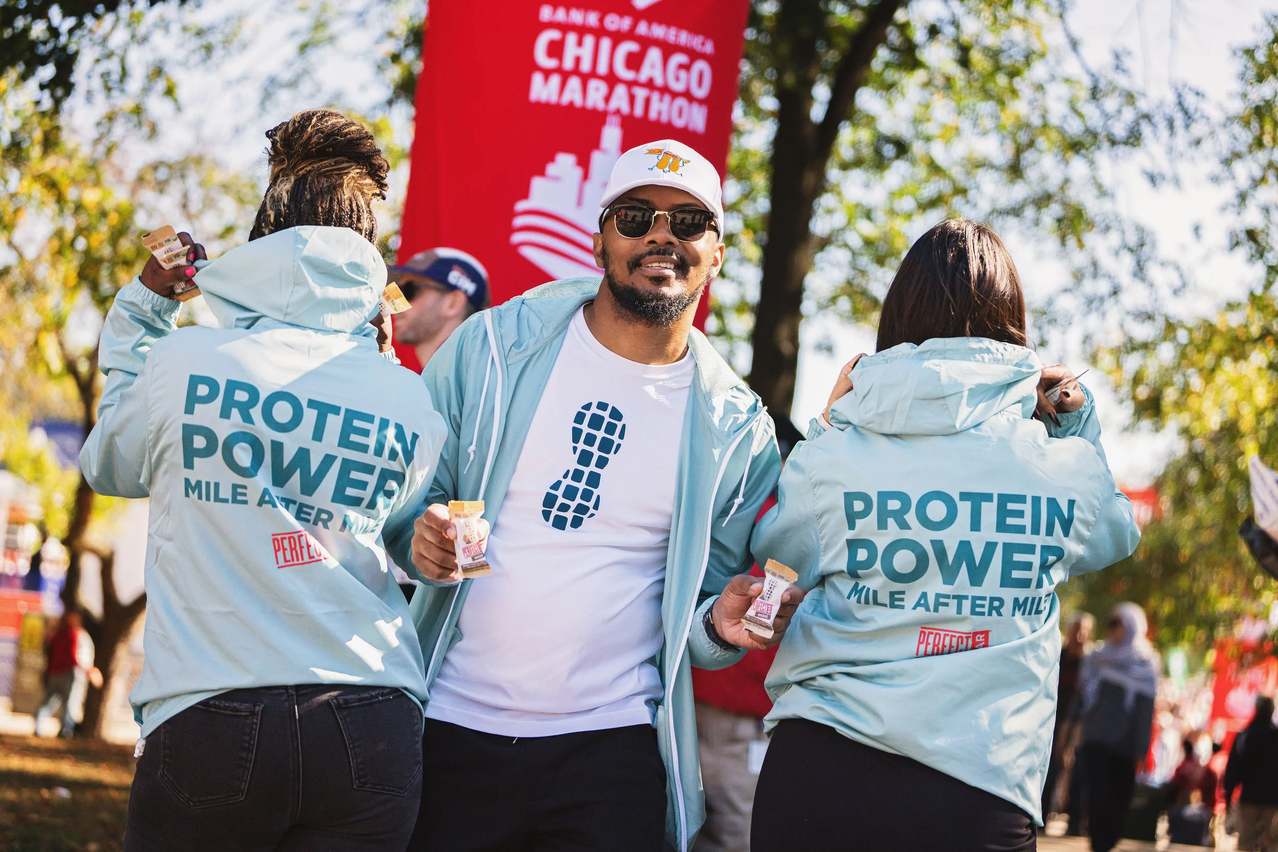 A man in a white shirt with a logo, wearing sunglasses and a cap, stands among event staff at the Chicago Marathon, with a large red banner and trees in the background.