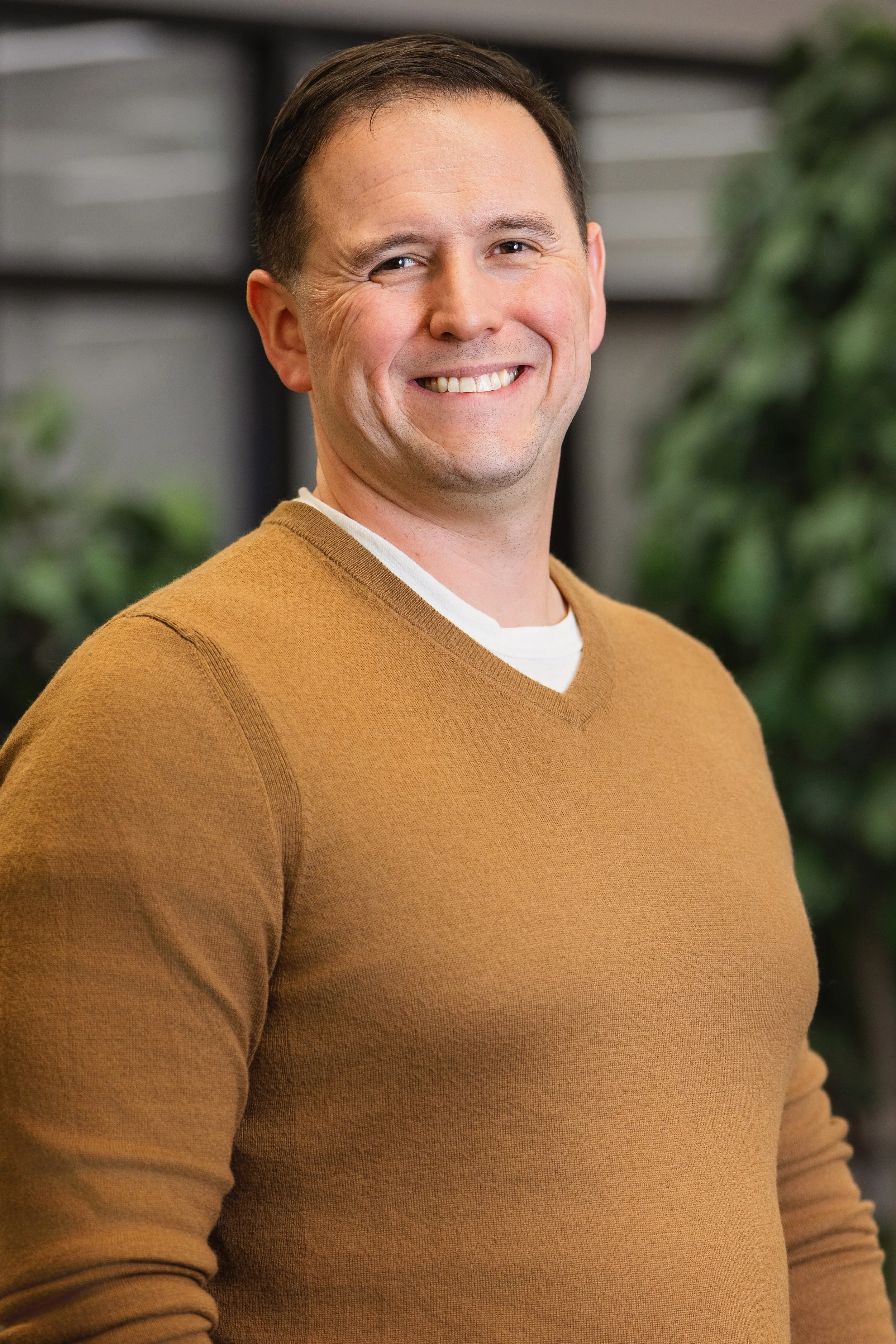 A smiling man wearing a tan sweater, standing in front of a blurred background with green plants.