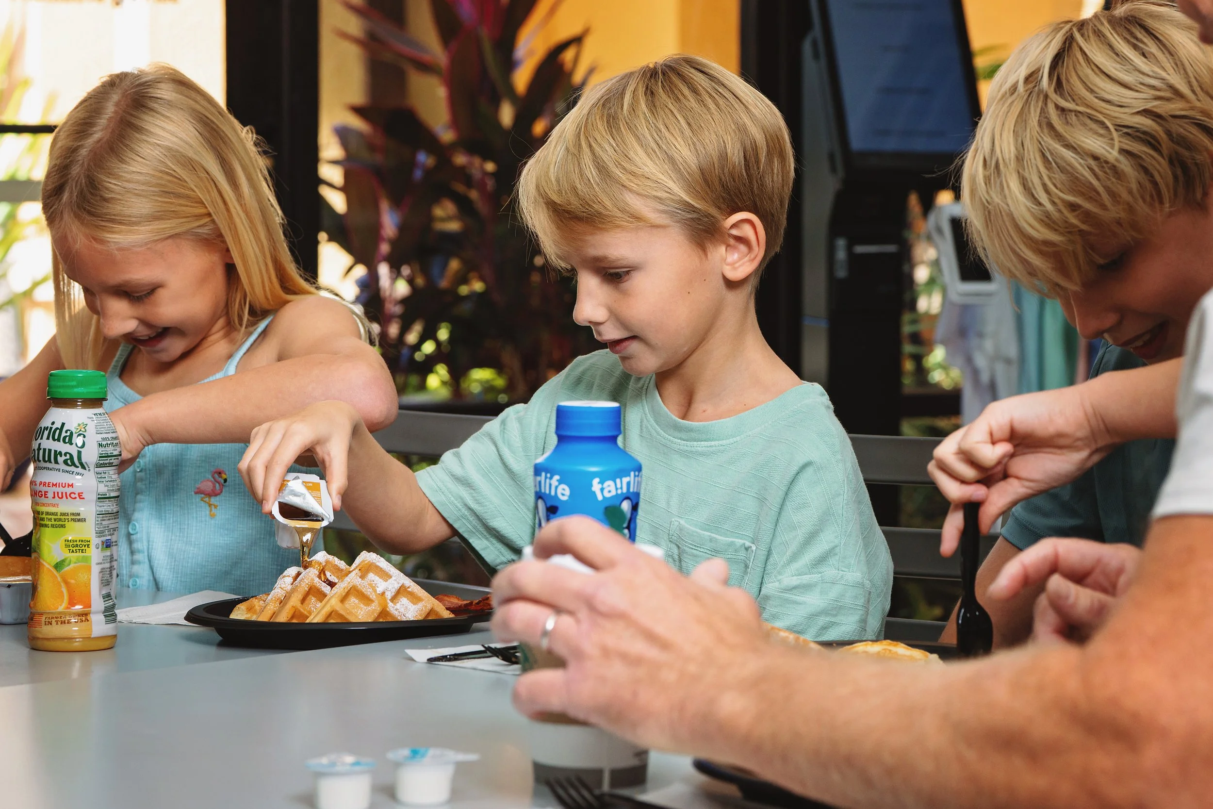 Children sitting at an outdoor table enjoying breakfast with waffles, orange juice, and yogurt, smiling and smiling.