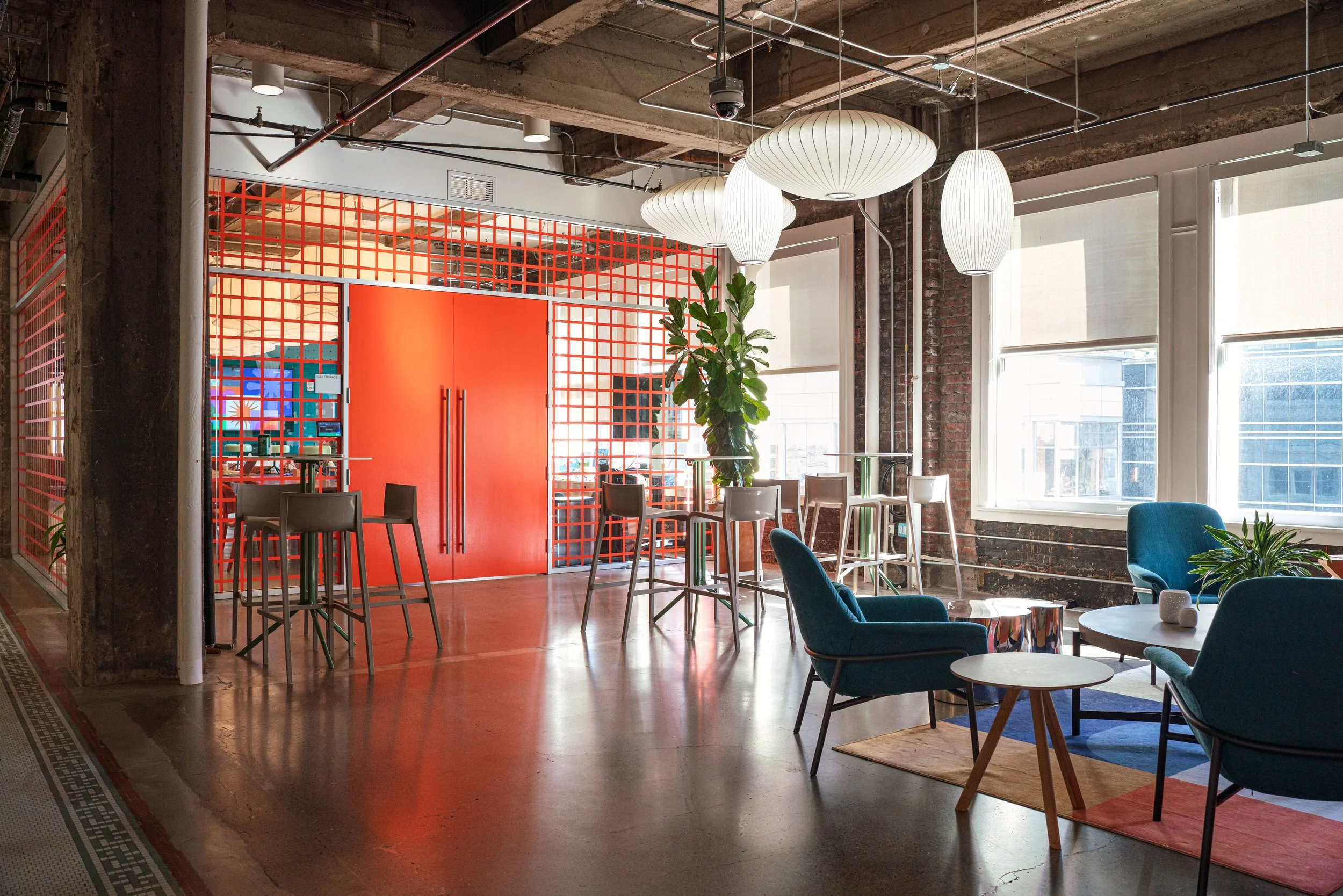 Modern open-concept lounge with bright lighting, large windows, colorful chairs, a potted plant, hanging paper lanterns, and a red lattice wall with a door.