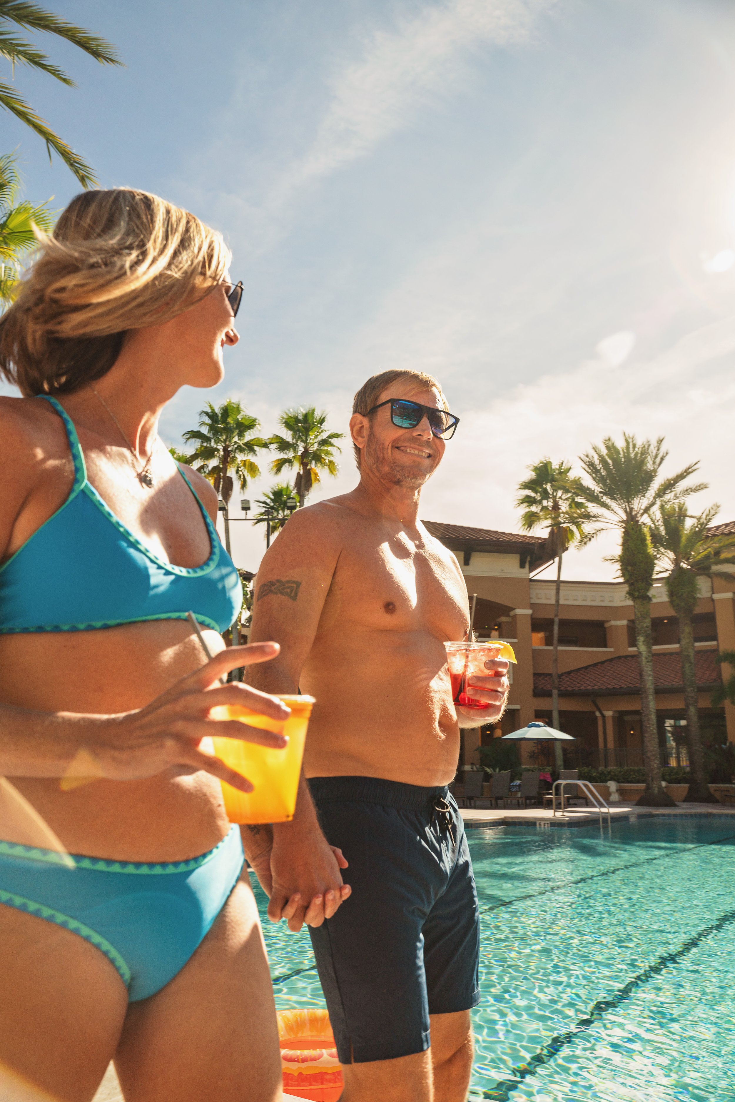 A man and woman in swimsuits standing by a pool, holding drinks and holding hands, with palm trees and a resort building in the background. It is sunny and the sky is mostly clear.