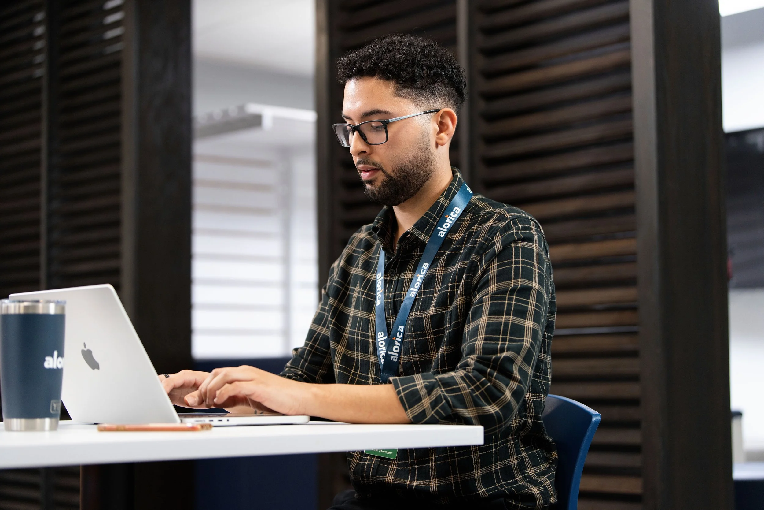 A man with glasses working on a laptop at a white desk in an office. He is wearing a green plaid shirt and a blue lanyard that reads 'alorica'. There are a stainless steel tumbler and a smartphone on the desk.