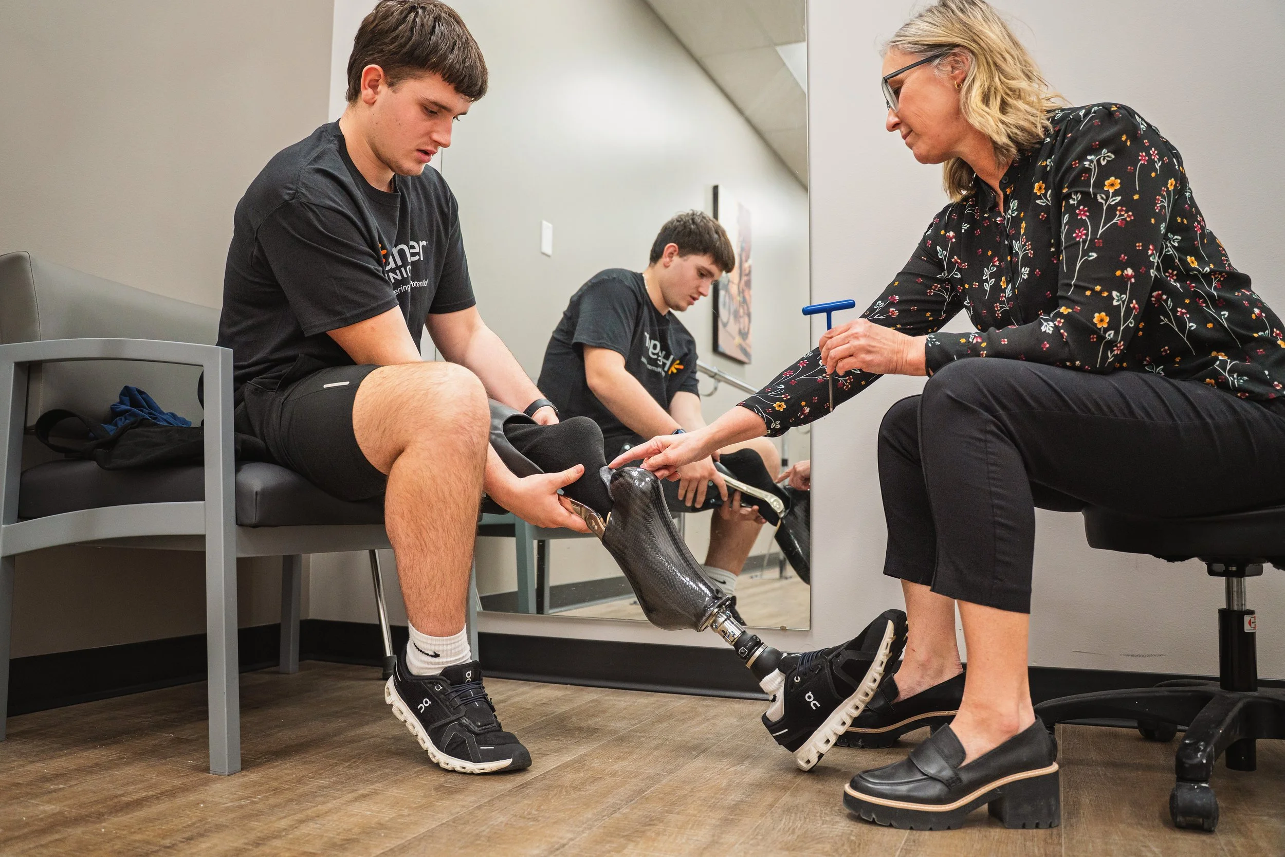 A healthcare professional adjusts a prosthetic leg for a young man sitting on a chair in a clinical setting while another young man waits in the background.