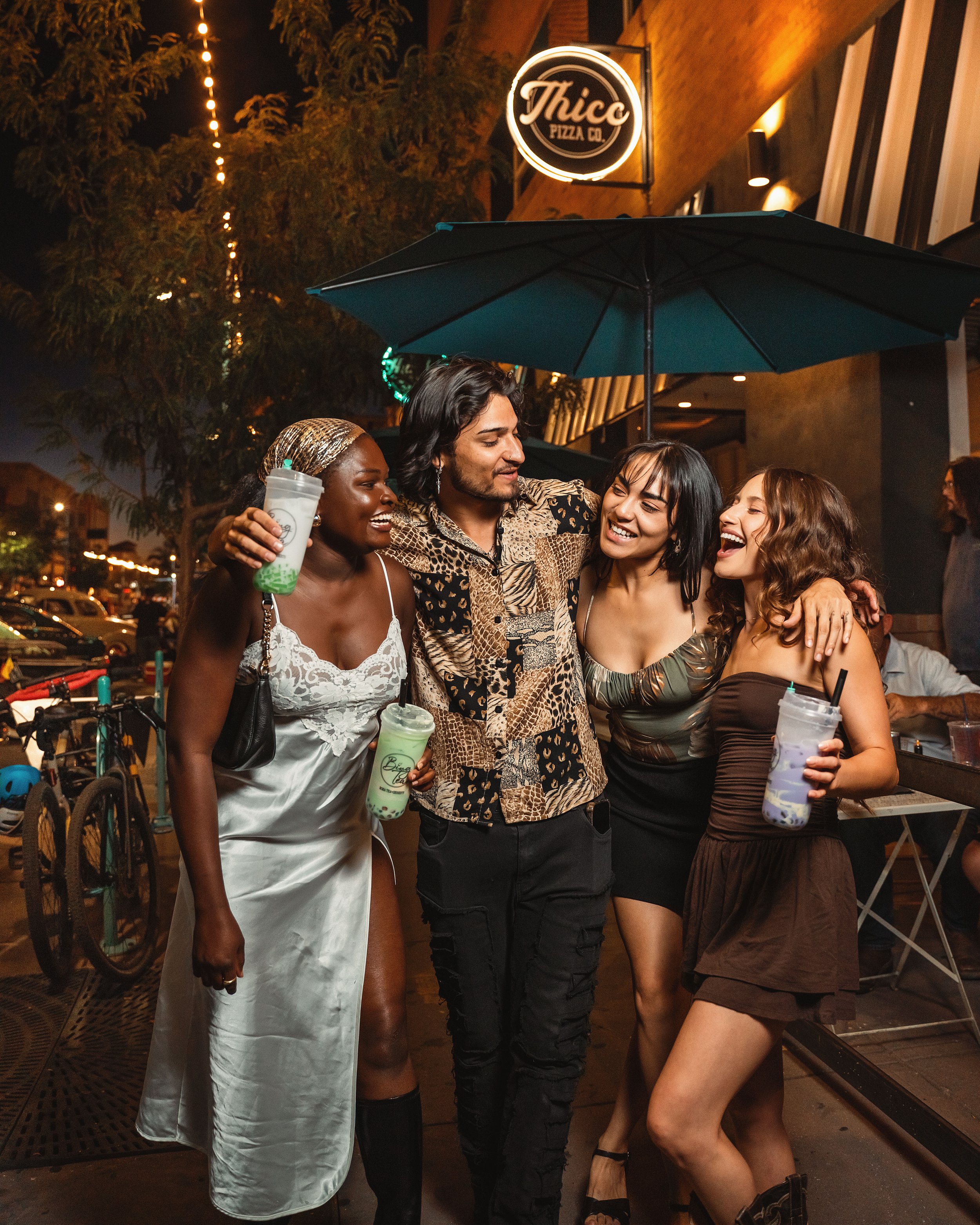 Four friends stand together on a city sidewalk at night, smiling and holding drinks, with a restaurant sign reading 'Thico Pizza Co.' above them.
