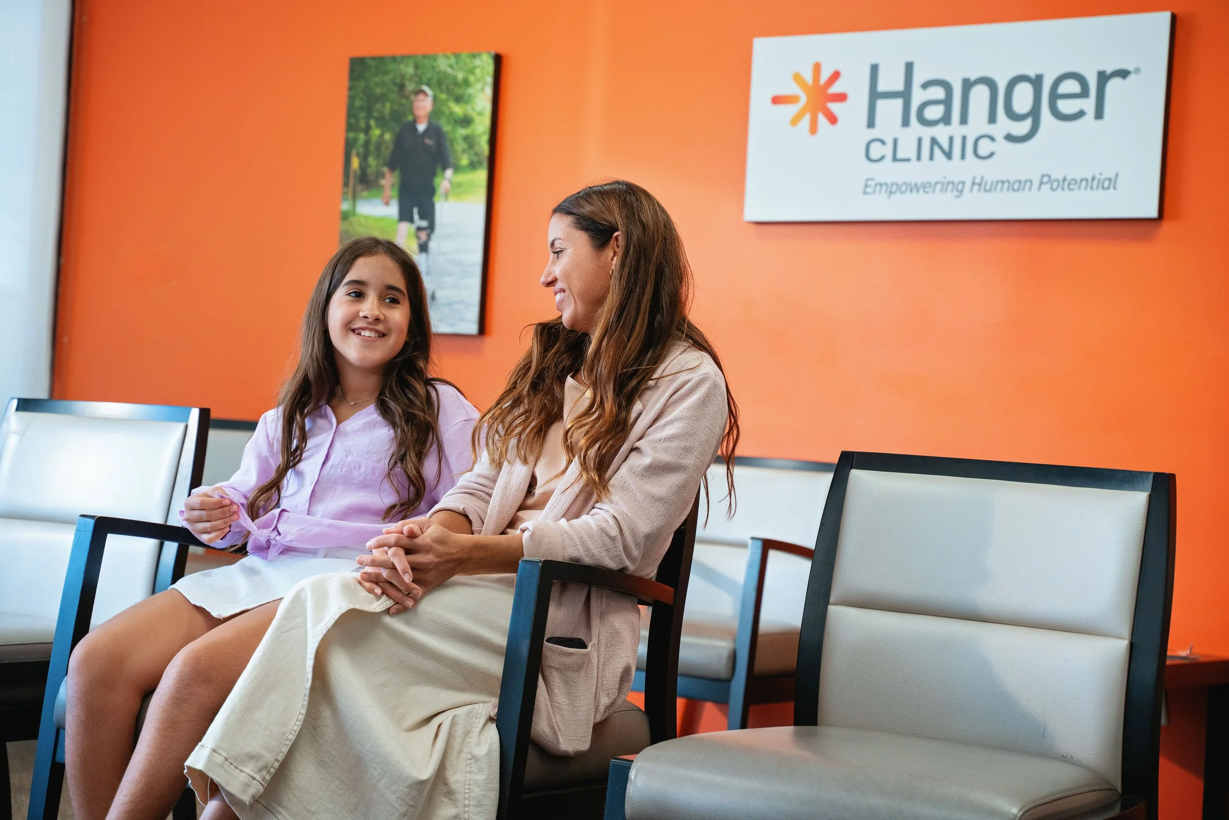 A woman and a young girl smiling and talking while sitting in a waiting room with chairs, in front of an orange wall with a Hanger Clinic sign.