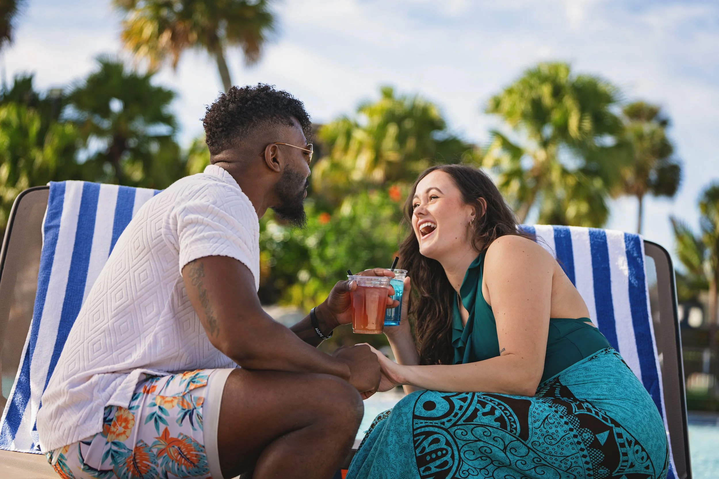 A man and woman with drinks are sitting in pool chairs at a poolside, smiling and engaging with each other with palm trees in the background.