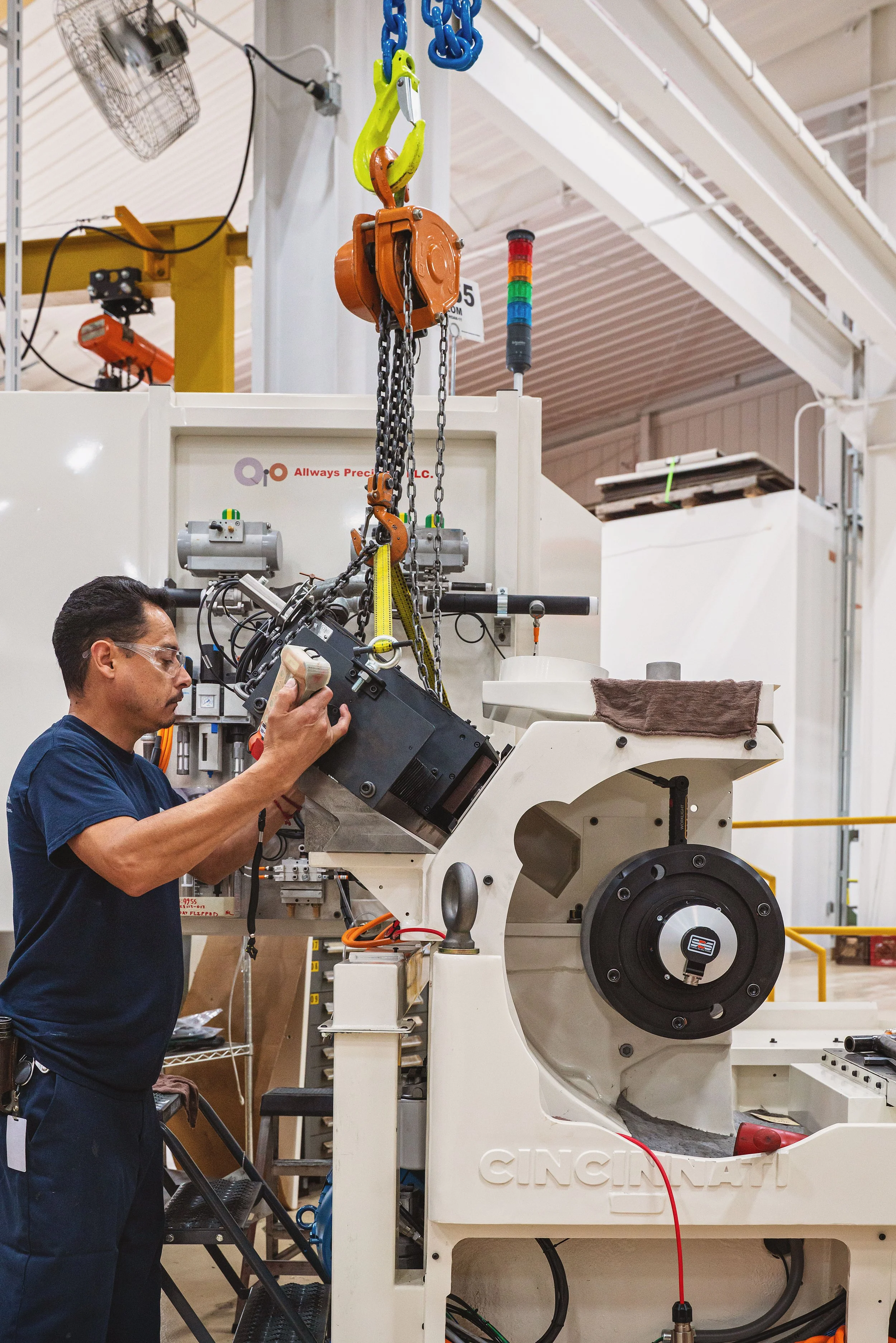 A man working with machinery in an industrial setting, surrounded by equipment and tools, with a large white machine labeled 'CINCINNATI' and a colorful signal tower in the background.