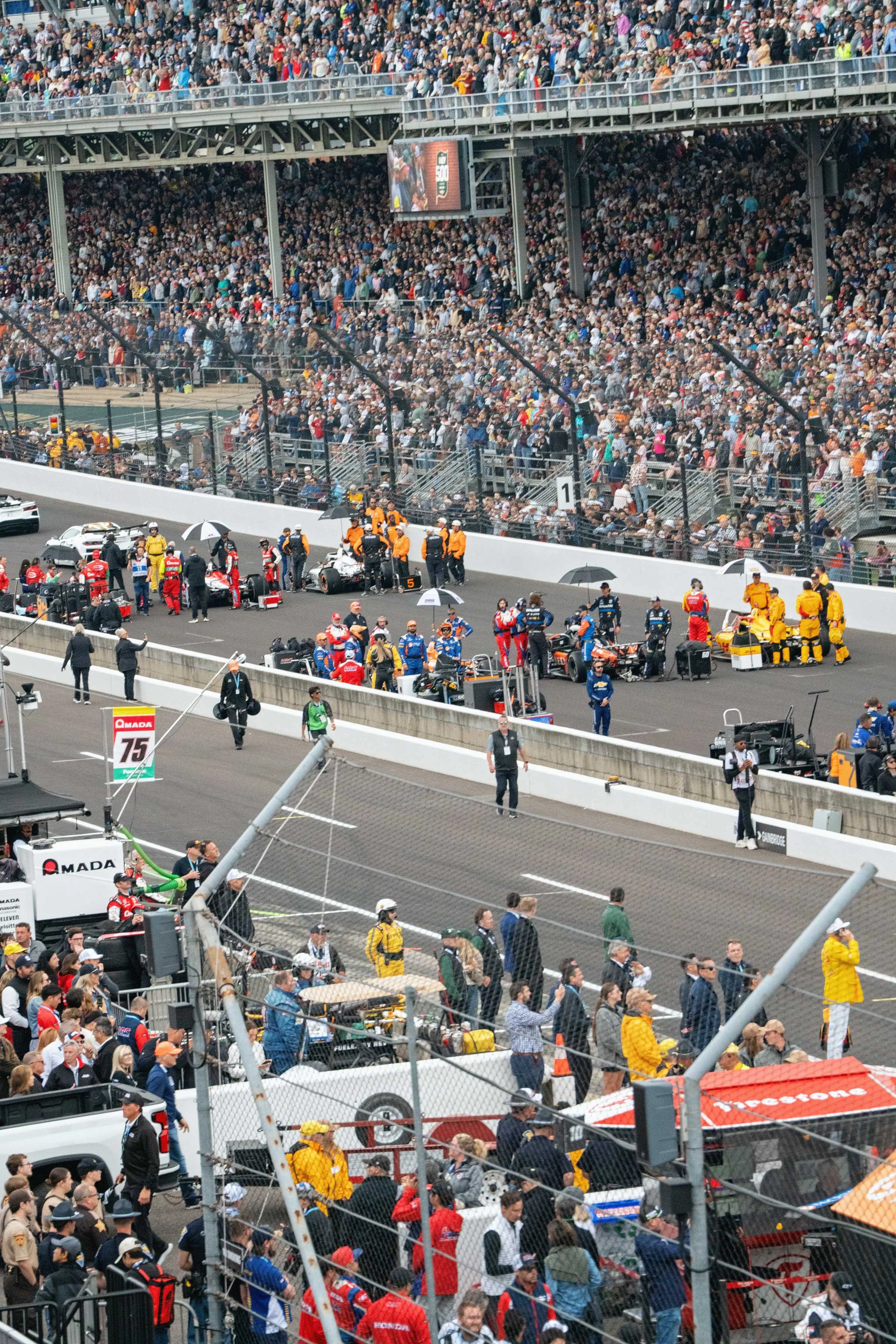 View of a crowded racetrack with racing cars, teams, and officials preparing for a race, surrounded by spectators in the grandstands.
