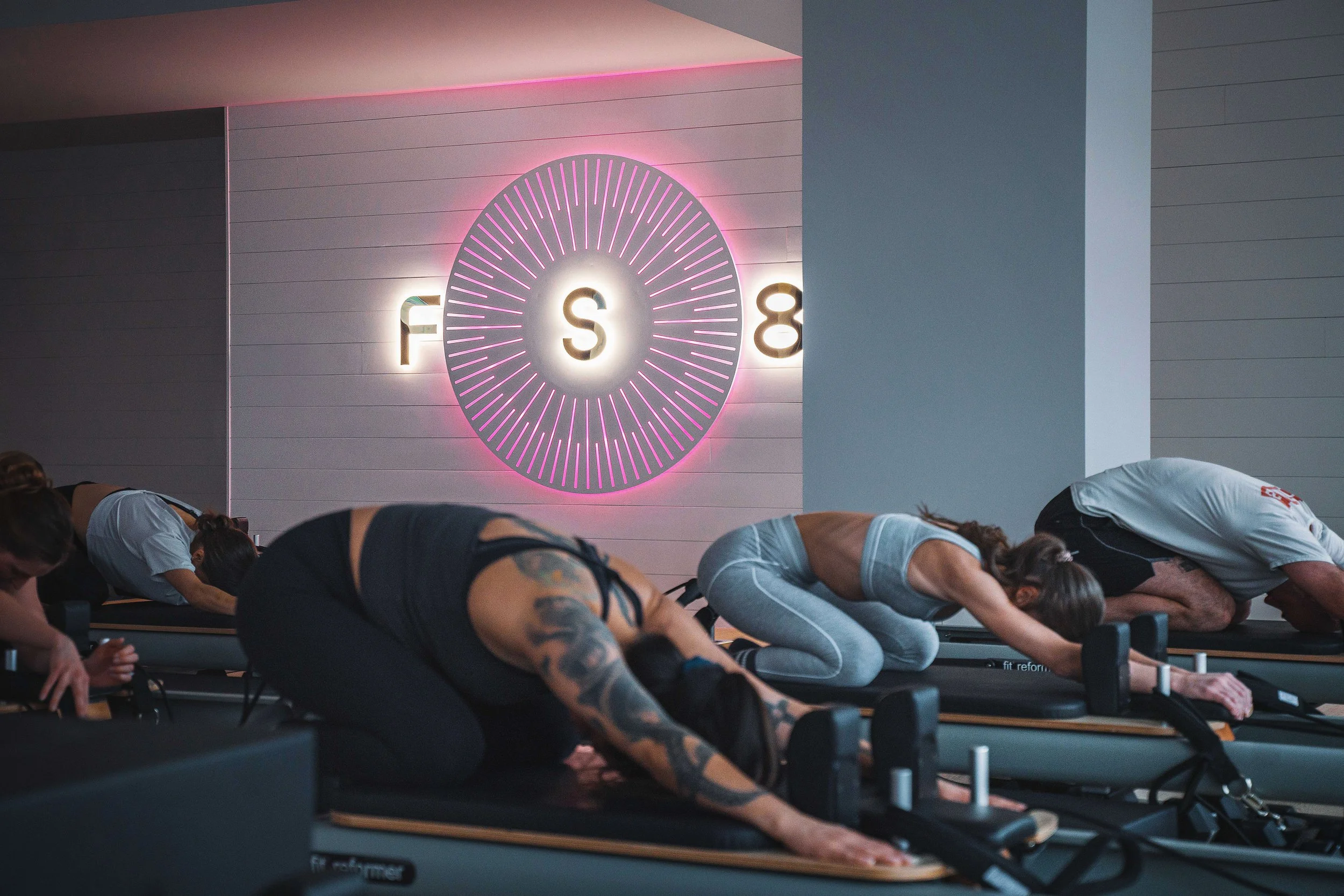 People participating in a Pilates class on reformer machines in a modern fitness studio with a neon sign on the wall.
